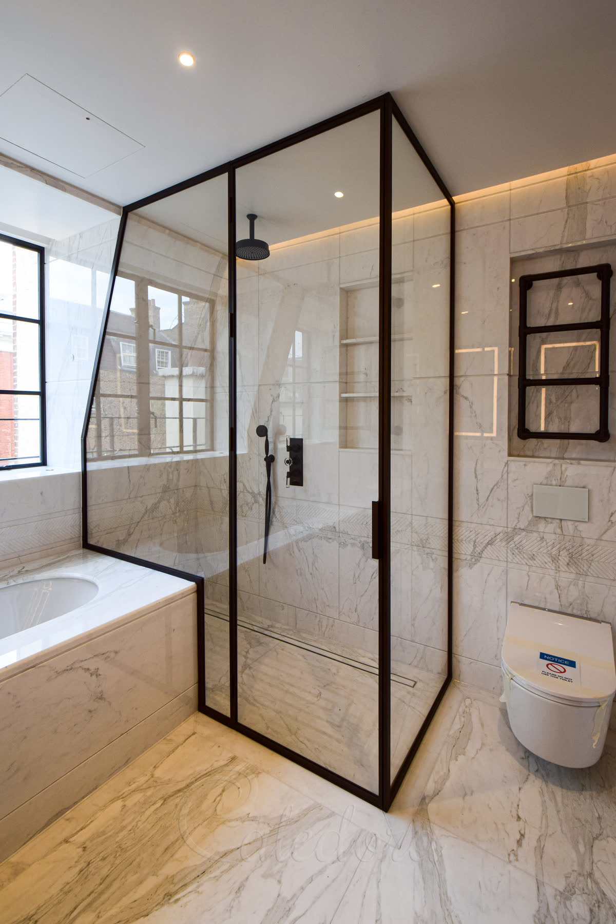 Bronze framed shower enclosure fitted over bath with custom glass cut following the bath's contour in a marble bathroom