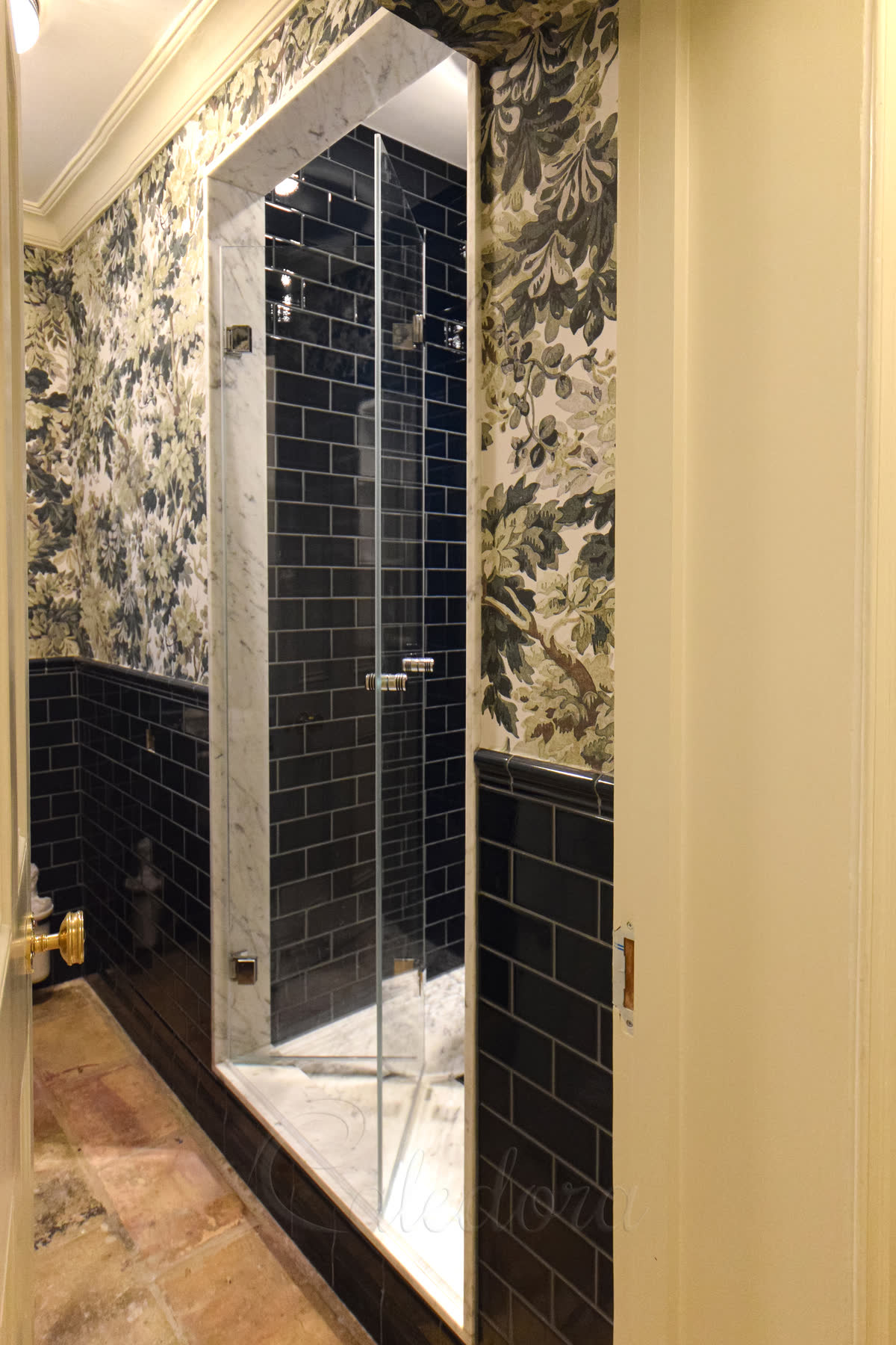 Frameless bifold shower door fitted to a very narrow alcove — black subway tile inside the enclosure and floral wallpaper either side, opening too tight for either inward or outward hinged swing so the bifold folds flat in place