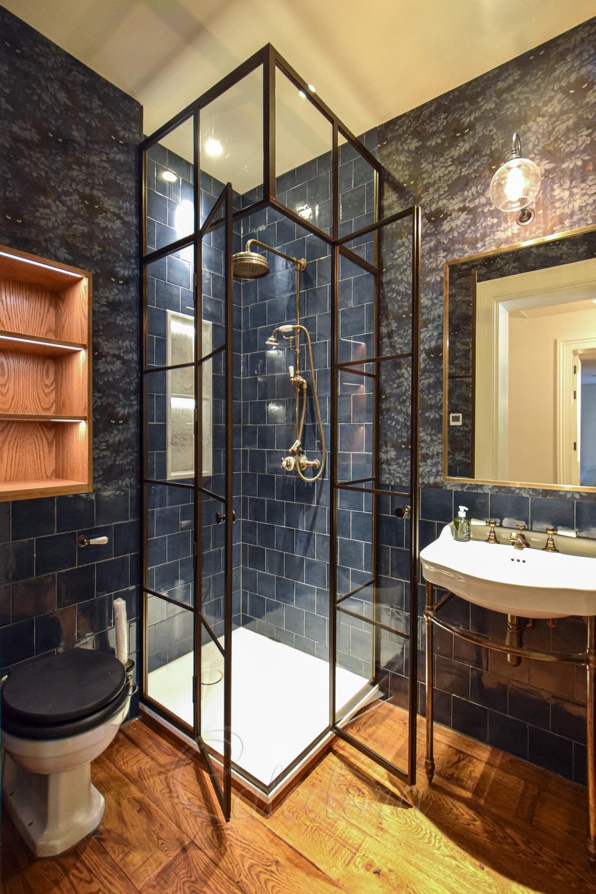 Bronze powder-coated framed corner enclosure doors with grid pattern and ultra clear glass — corner-entry layout fitting between a toilet and vanity, brass shower controls inside