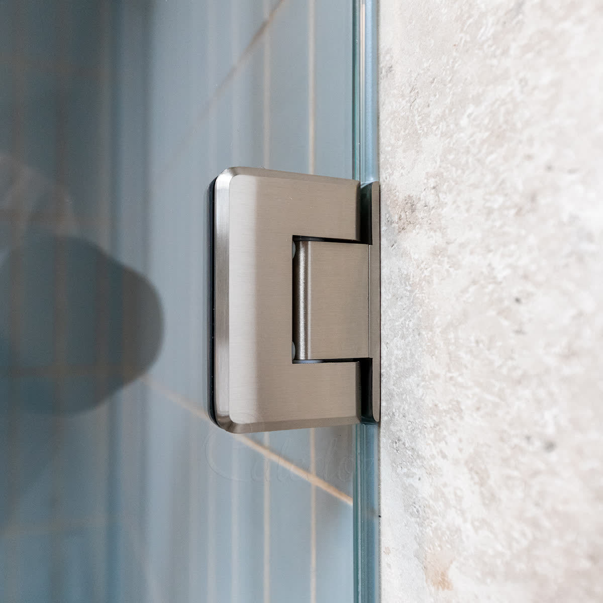 Close-up of a brushed nickel frameless hinge fixed to a travertine wall — the off-set plate's smooth bathroom-facing face shows no screw heads, the fixings sit behind it (Caledora's standard back-screw configuration)