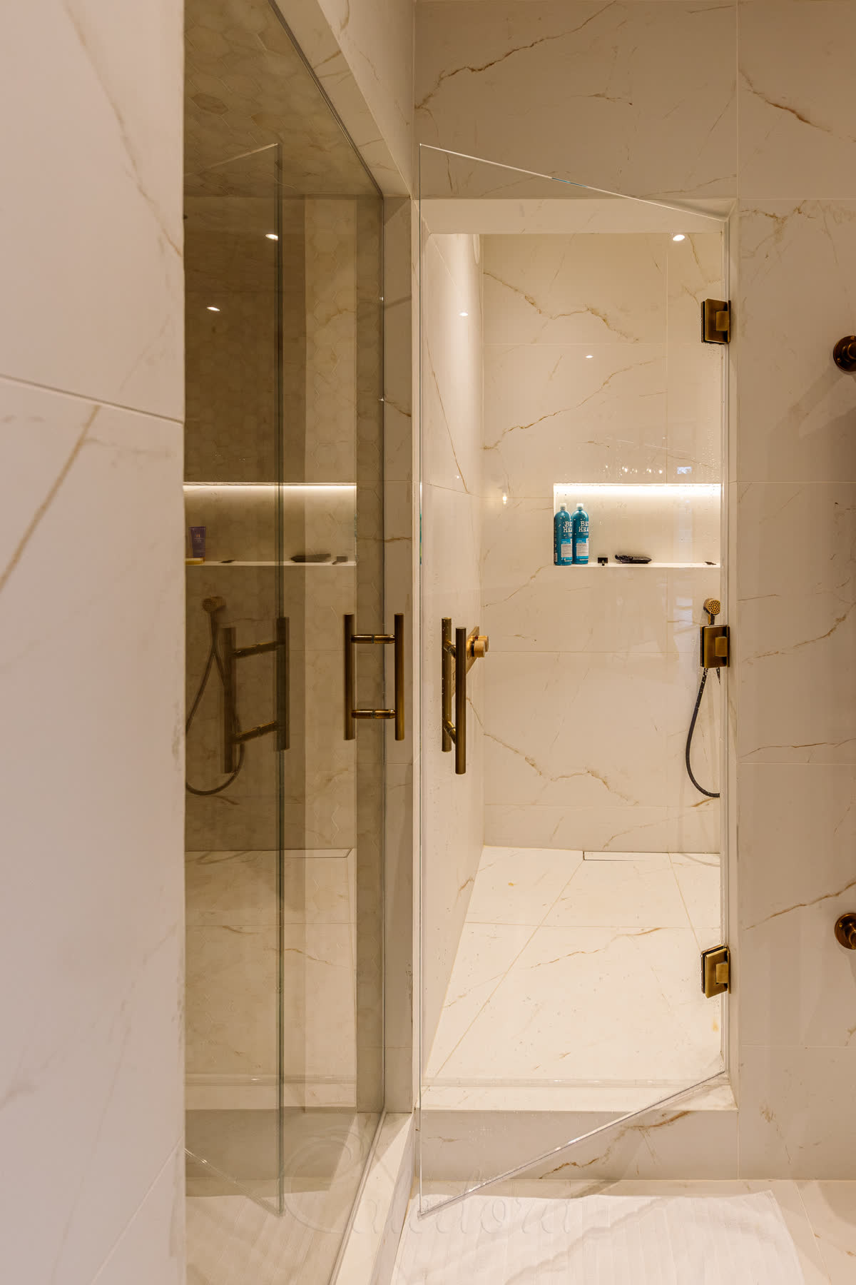 Antique brass swing shower door opening inward, three hinges clear wall-mounted brass shower controls — coordinated with a matching steam-room door and sauna door in the same suite