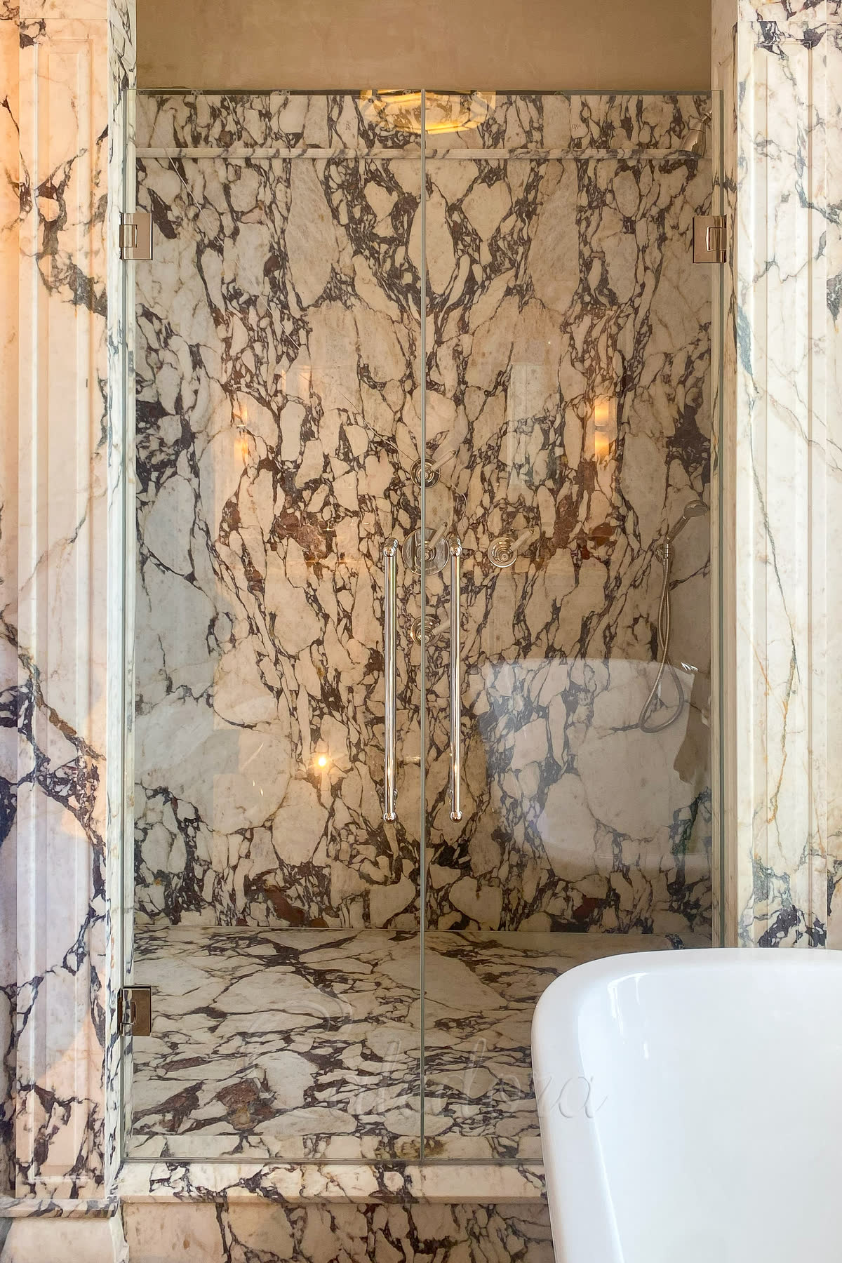 Two frameless hinged shower doors meeting in the middle against a wall of dramatic Calacatta Viola marble — brushed brass T-bar handles, brushed brass hinges, wall sconces either side and a freestanding tub partially visible