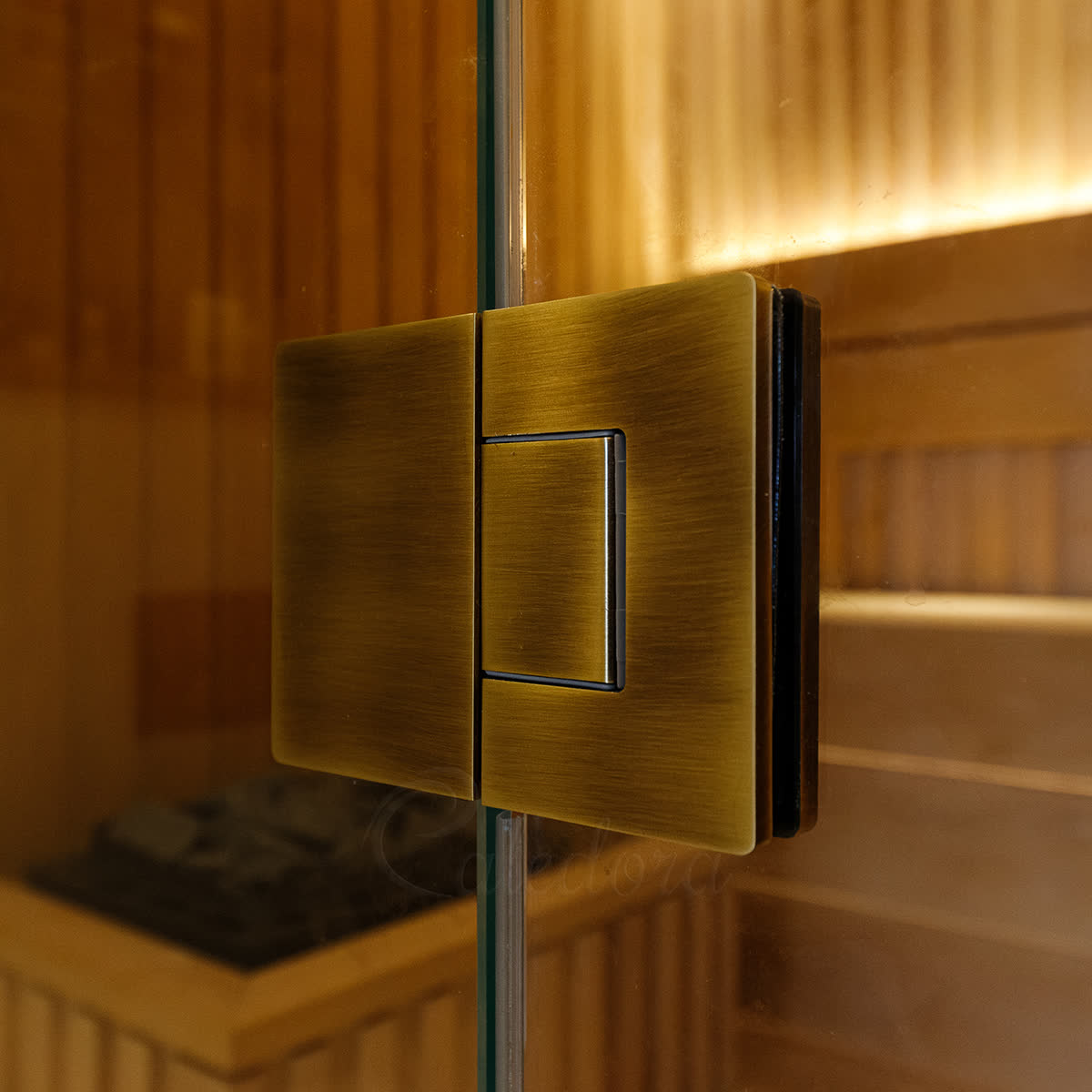 Close-up of an antique brass glass-to-glass frameless hinge — two matching plates clamping a fixed glass panel and the door glass, no wall behind, demonstrating the glass-to-glass mounting style