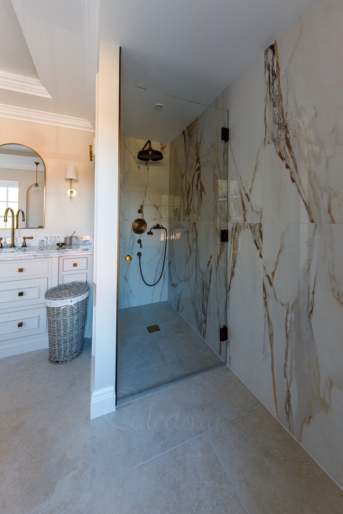 Frameless hinged shower door with three antique brass hinges mounted directly into a Calacatta marble wall — Glass-to-Wall configuration, closed-position view from outside the enclosure showing the wider bathroom with white vanity unit