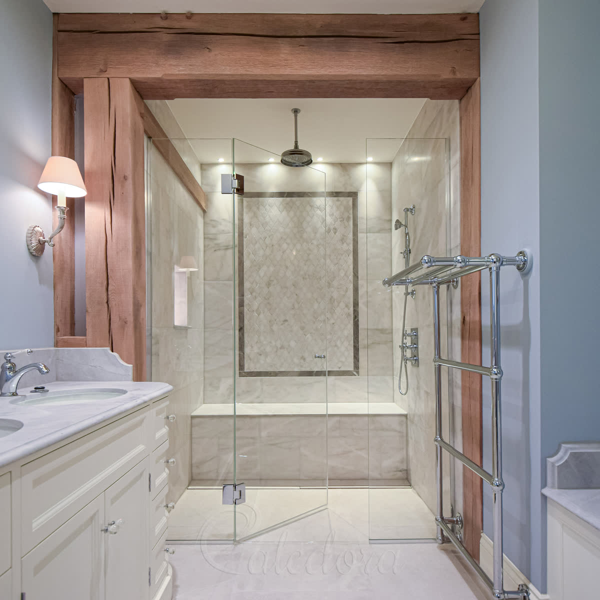 Frameless inline hinged shower door with polished chrome hardware on a marble wall — door and fixed glass panel sit in the same plane at 180°, herringbone marble feature panel inside the enclosure, heritage chrome towel radiator alongside and a rustic timber frame around the opening