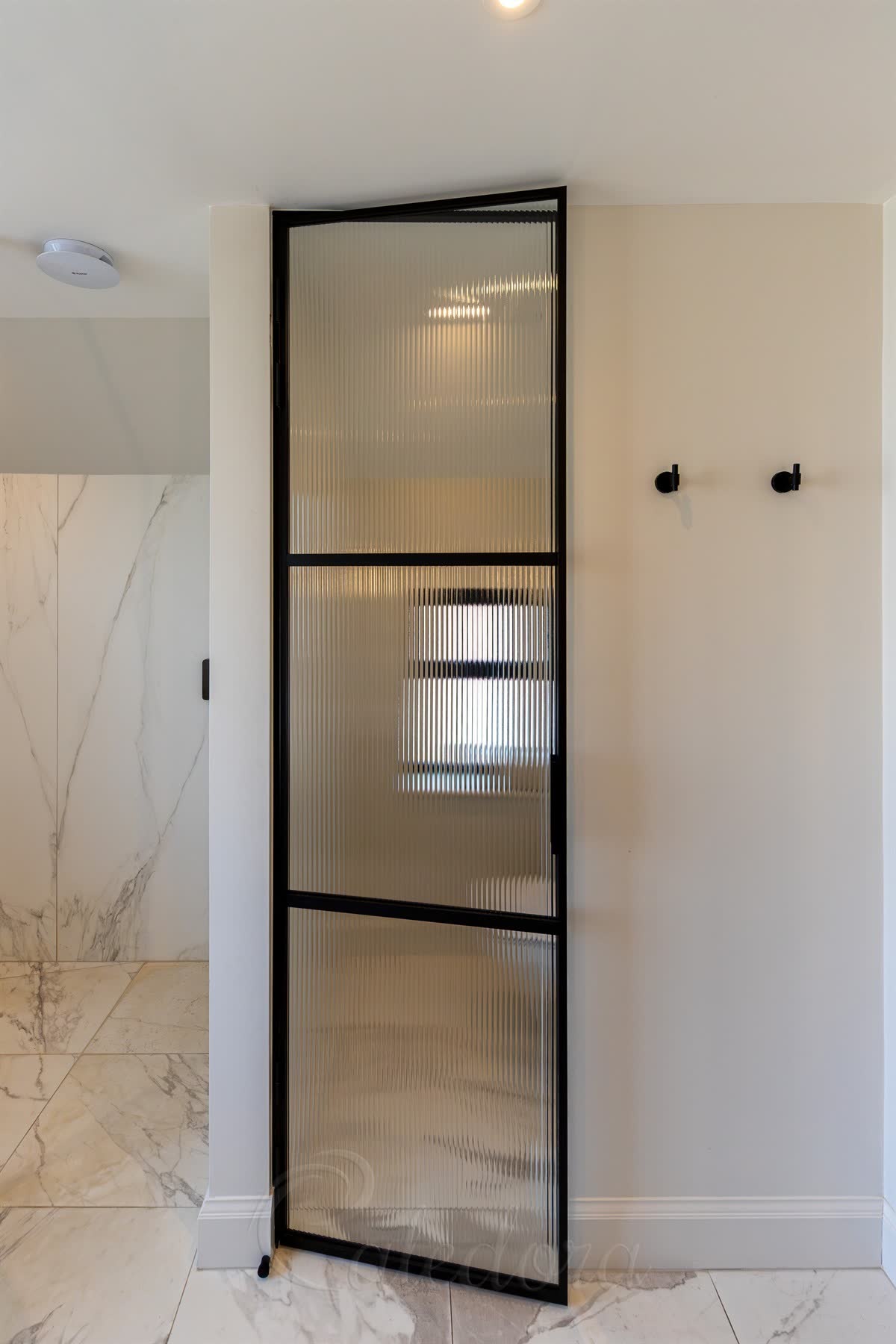 Black framed shower door with reeded laminated glass — slim three-rail matt black frame standing ajar against cream walls, with veined marble floor and wall beyond