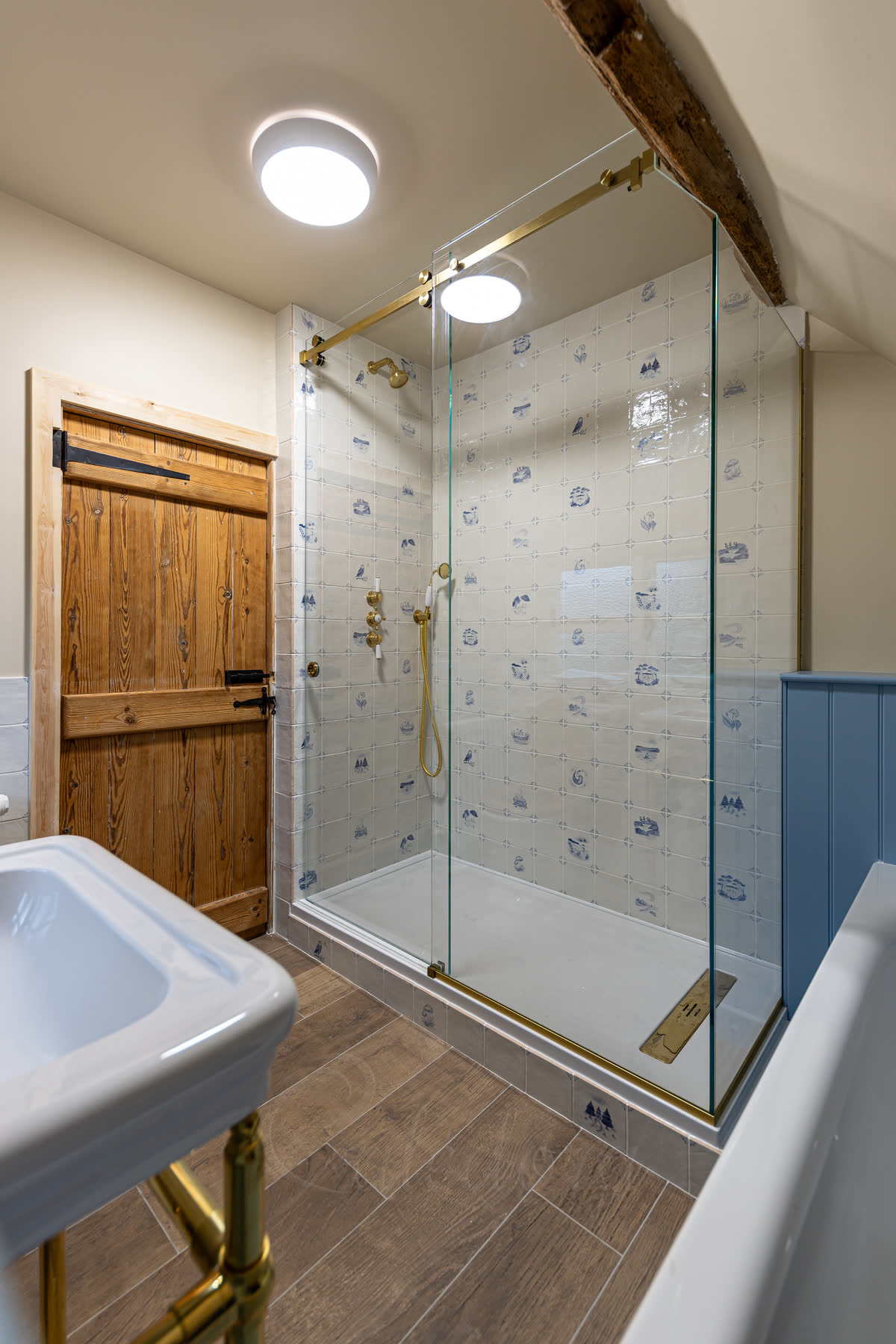 Frameless sliding shower door with brushed brass track in a nautical-themed bathroom with exposed wooden beam ceiling and matching brass shower fixtures
