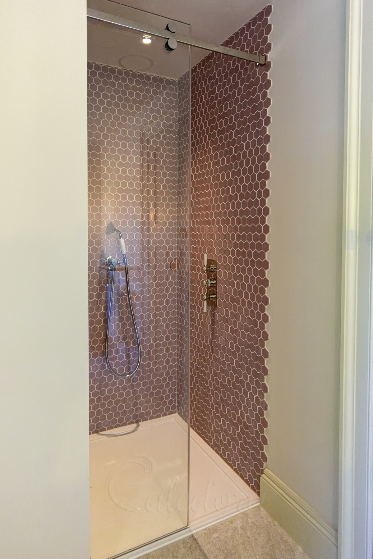 Glass sliding shower door with chrome track sliding behind a wall, in a bathroom with pink hexagonal mosaic tile walls and brass shower fixtures