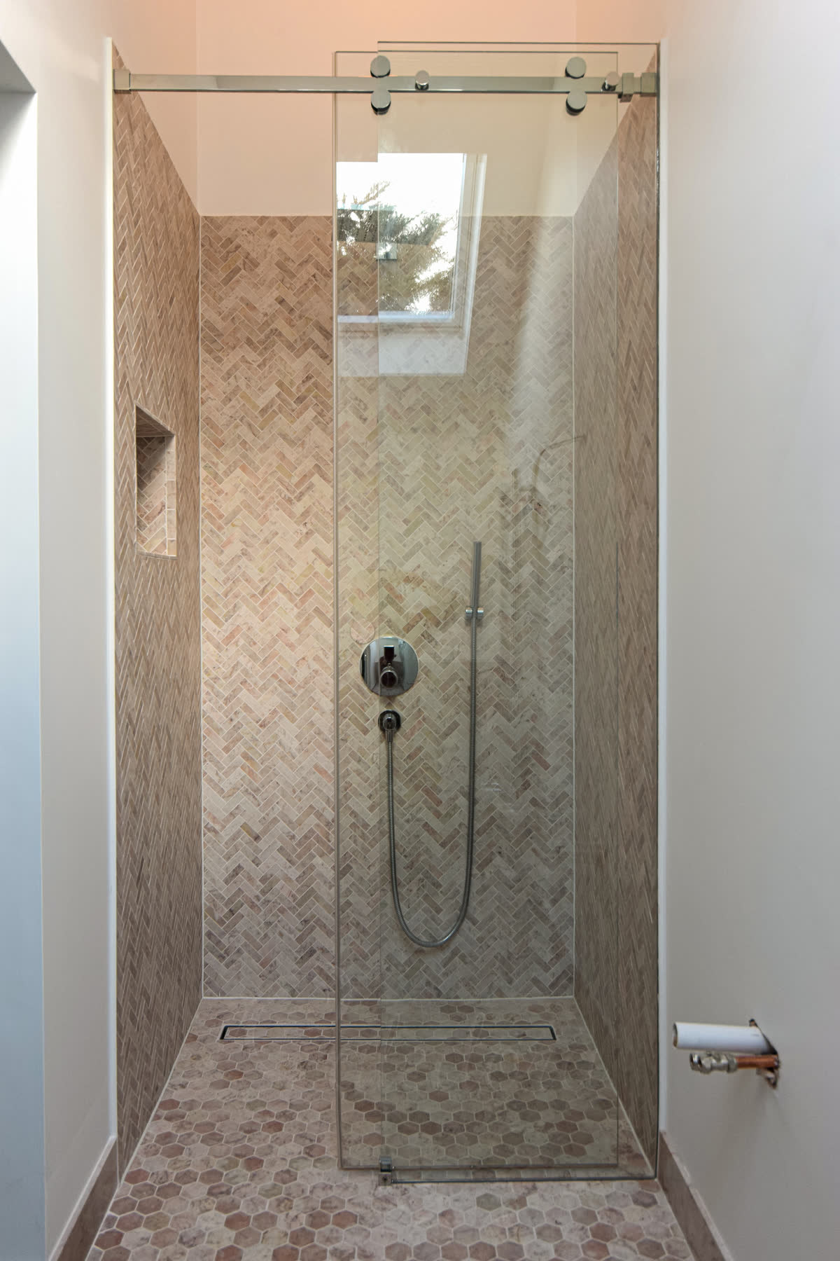 Compact frameless sliding shower door with chrome track in a narrow enclosure with herringbone mosaic walls and hexagonal floor tiles