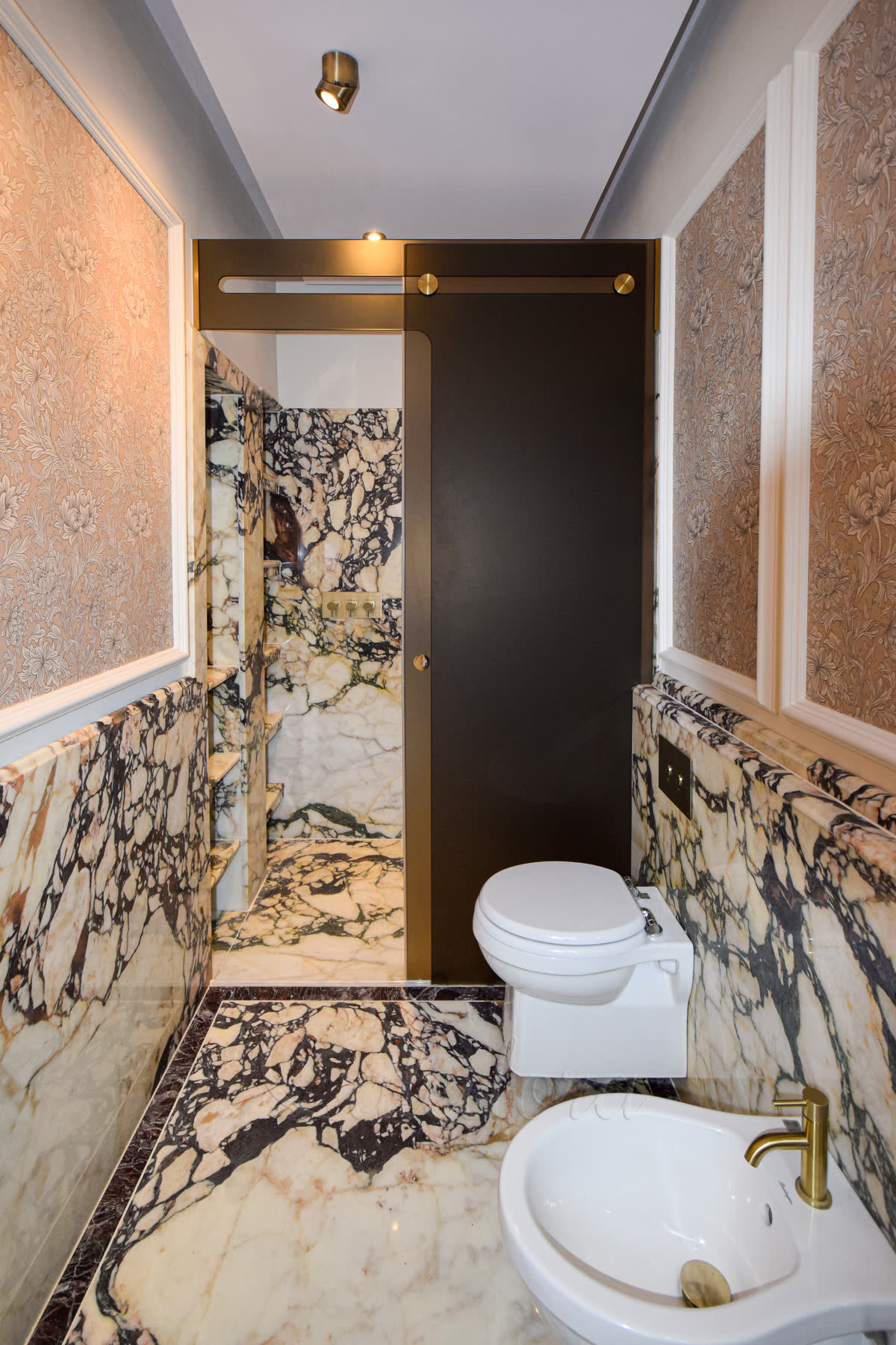 Trackless sliding shower screen with bronze tinted frosted glass and a glass-supported door mechanism in a luxury bathroom with marble walls and brass fixtures