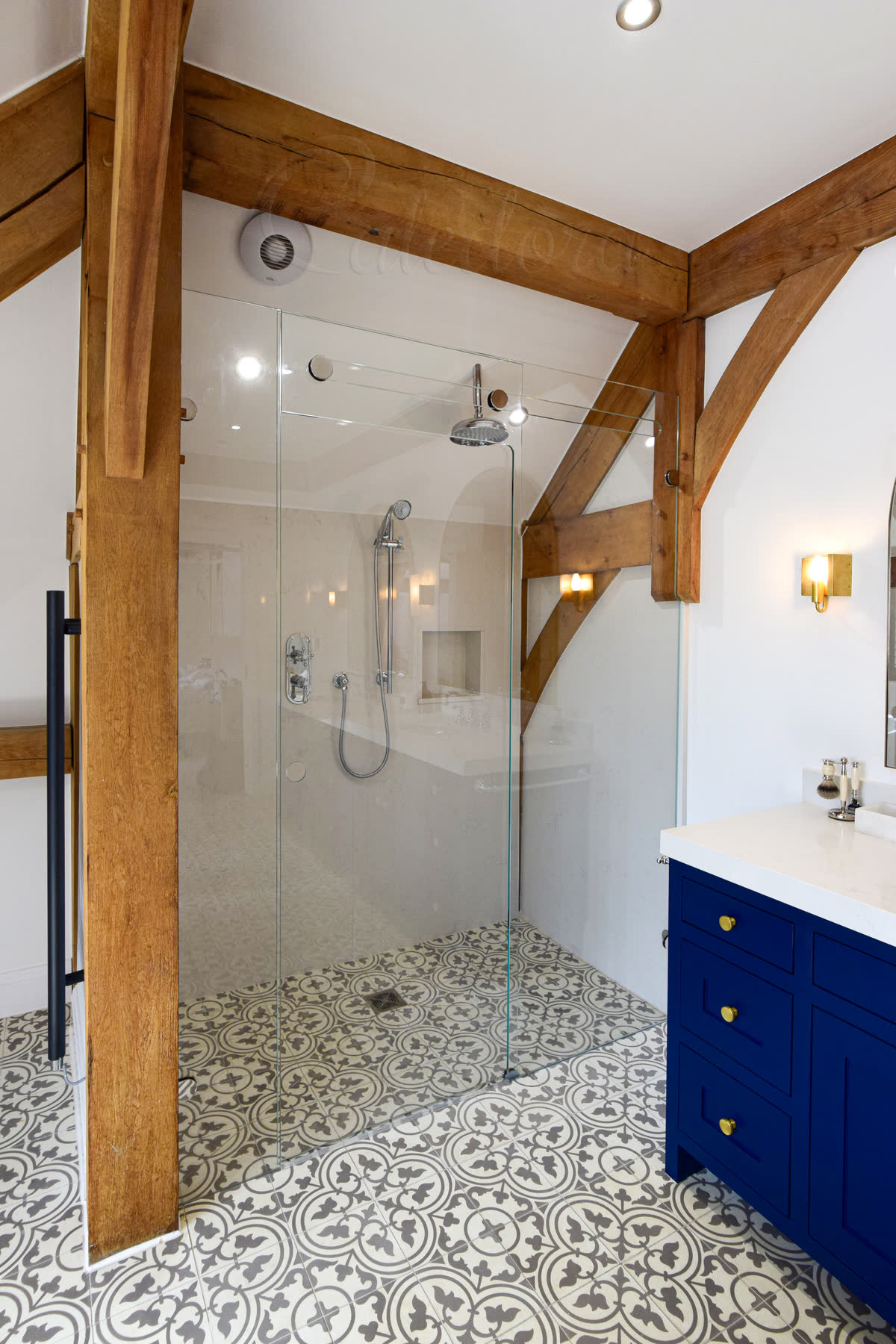 Trackless frameless sliding shower screen with glass-supported door mechanism, exposed oak beam ceiling, decorative patterned floor tiles and navy blue vanity