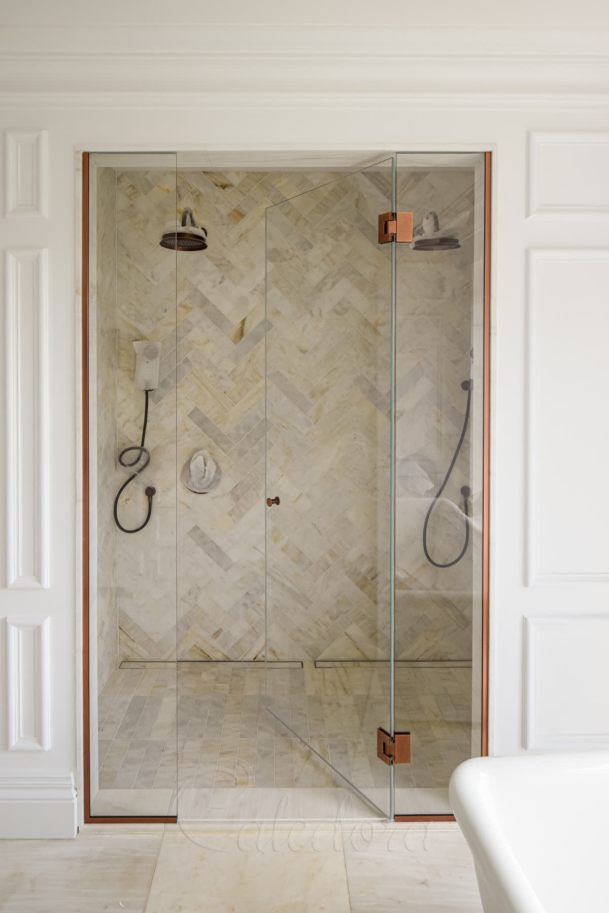 Frameless inline shower enclosure with antique copper hinges for two people, herringbone marble flooring