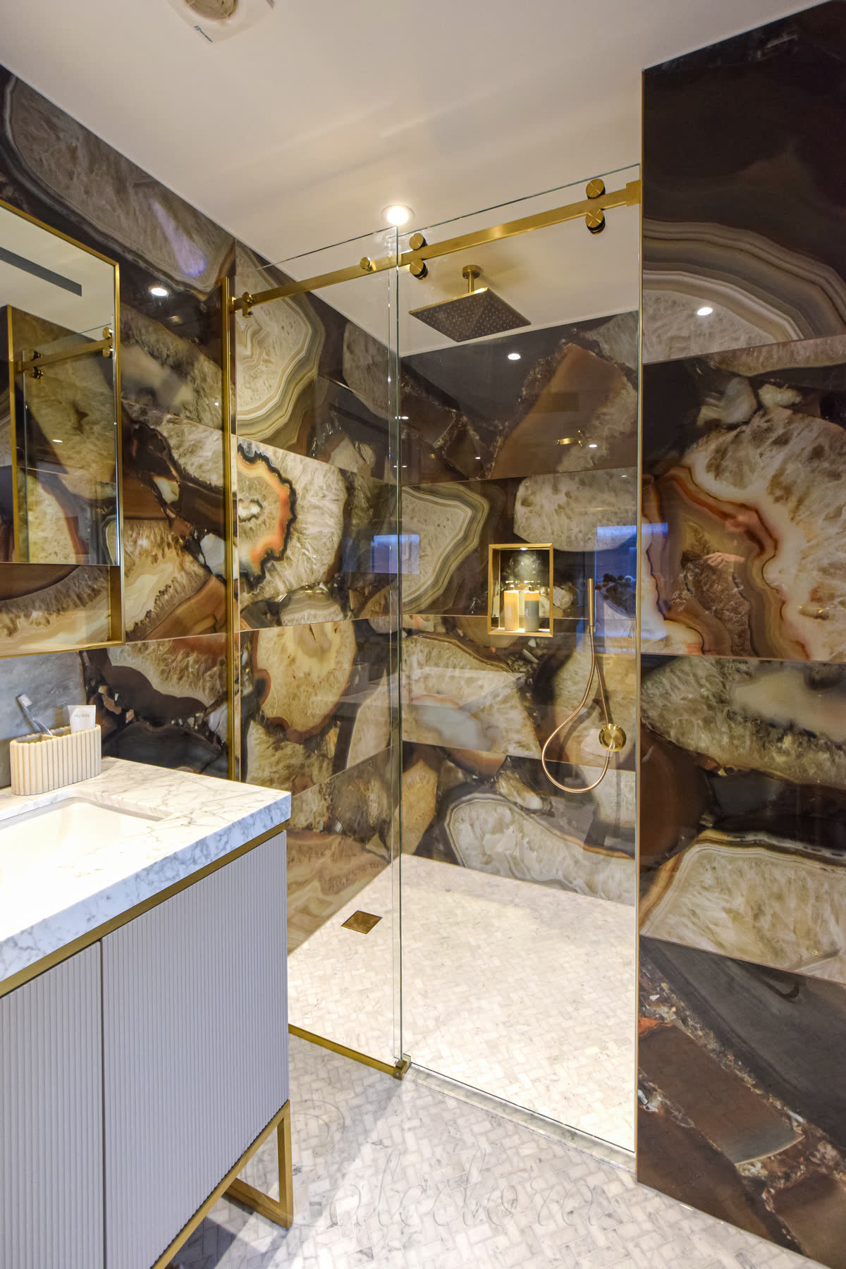 Frameless sliding shower door with brushed brass track system positioned in bathroom alcove featuring dramatic natural stone walls with gold veining and coordinating brass fixtures