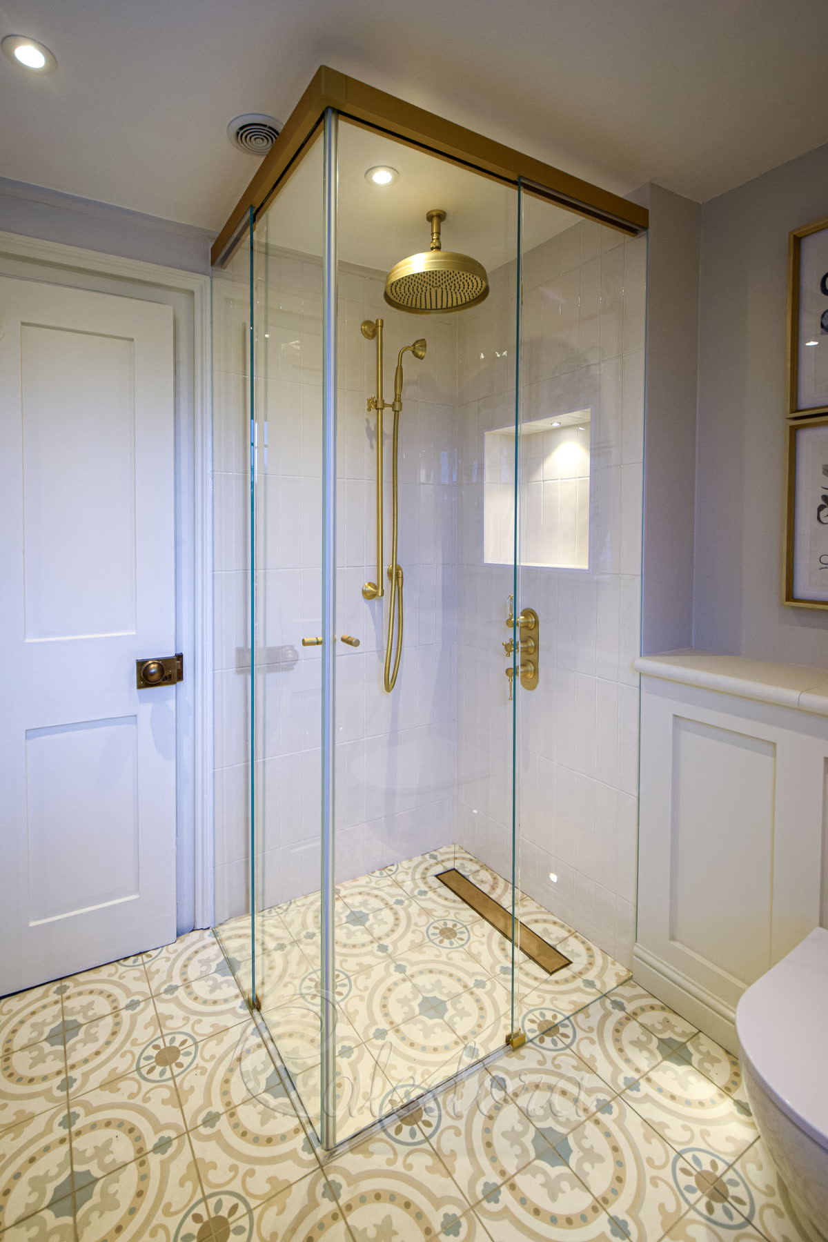 Corner sliding shower doors with brushed brass track system featuring double glass panels, coordinating brass rainfall showerhead and fixtures, positioned over ornate geometric patterned floor tiles