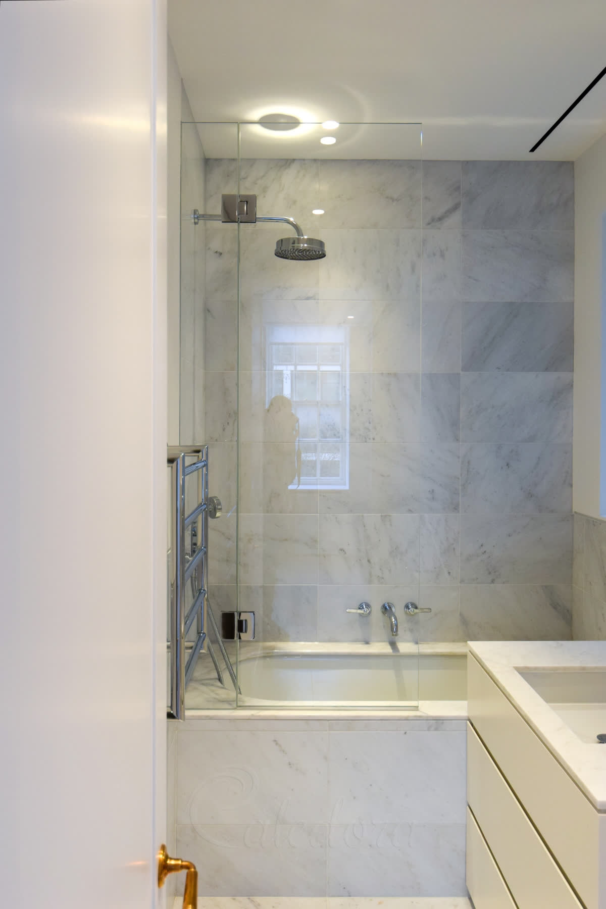 A bath screen with a fixed panel and a hinged panel beside a chrome heated towel rail, in a grey marble bathroom with a white vanity