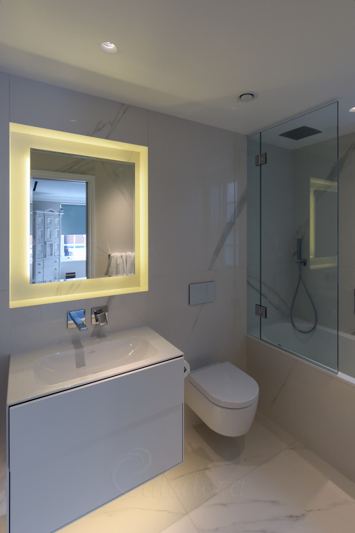 A frameless bath screen at the end of a bath beside a wall-hung toilet, with a backlit mirror and a white vanity in a marble bathroom