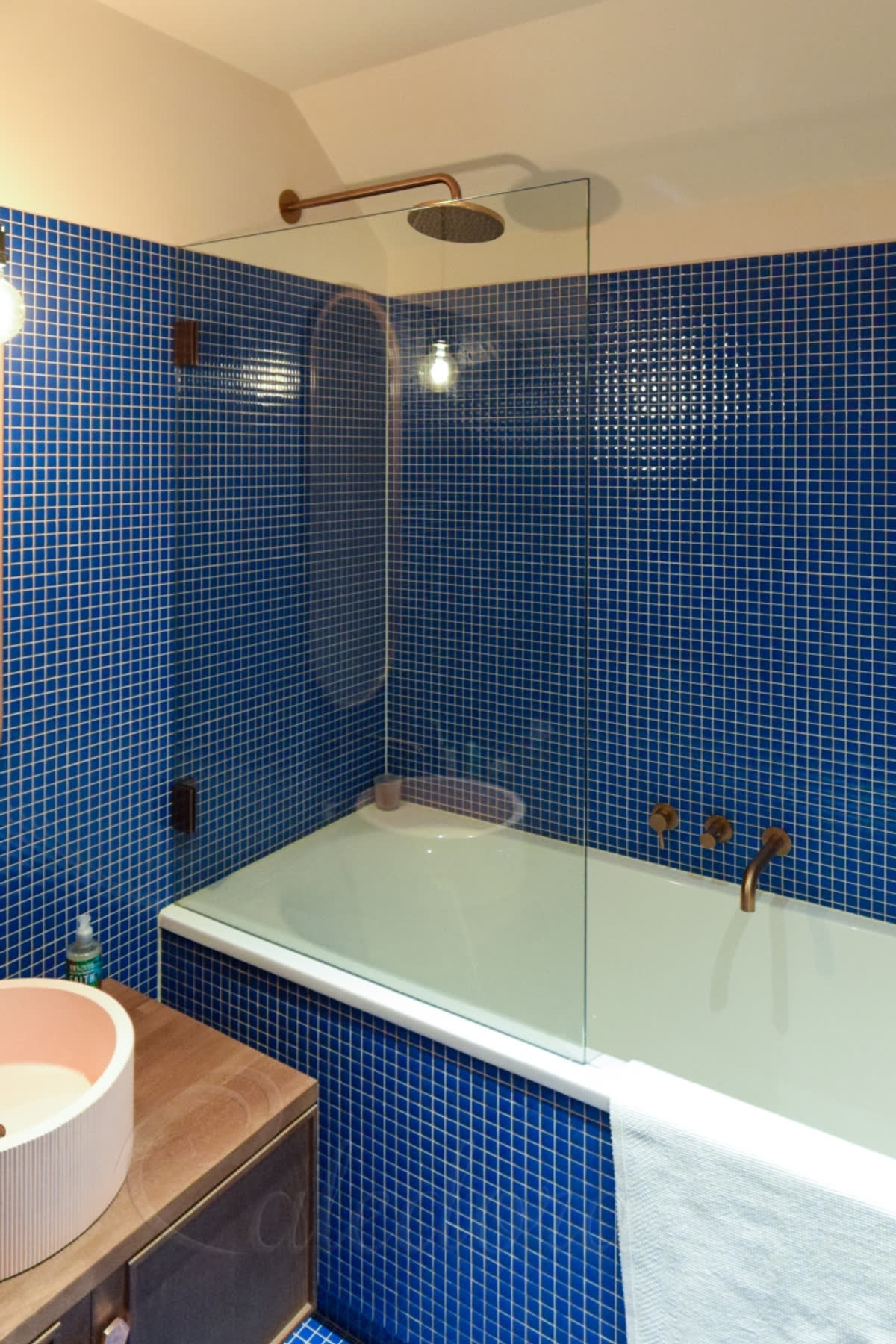 A glass bath screen hung on copper hinges at the wall over a bath in a blue mosaic bathroom, with a copper rain shower and taps