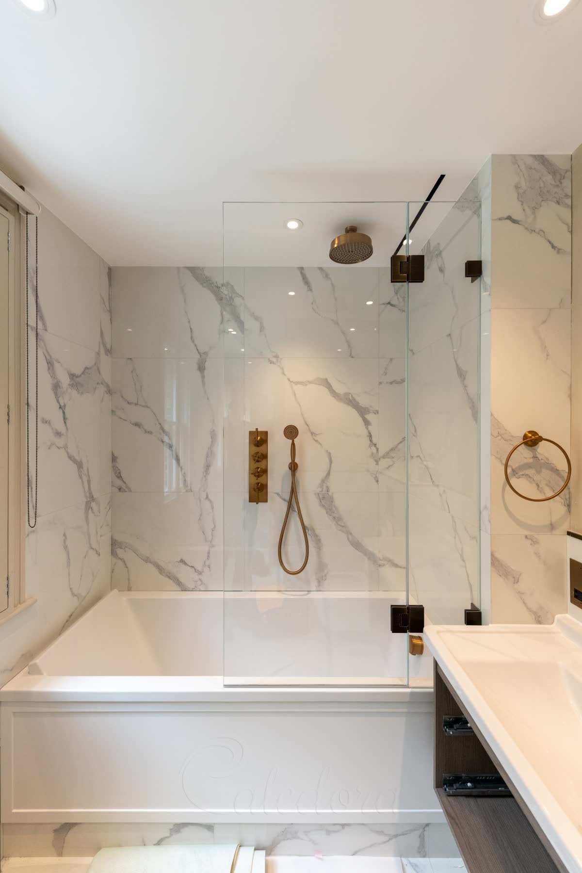 A bath screen with a fixed panel at the wall ending in line with the vanity unit beside the bath, its hinged panel on dark hinges, with brushed brass shower fittings and white marble walls