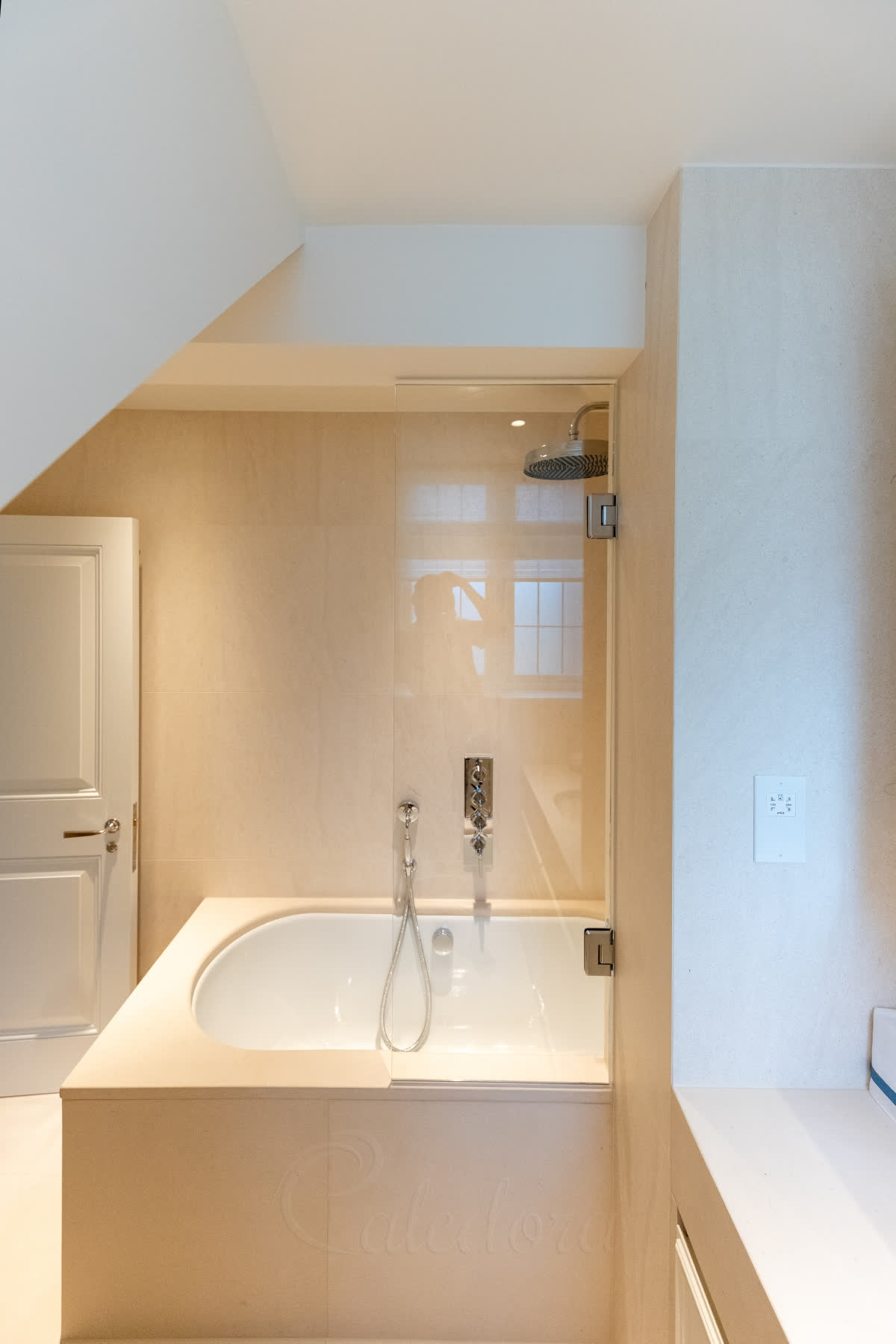 A rectangular glass bath screen hung from the wall on chrome hinges, over a stone-topped bath with chrome shower controls in a cream bathroom