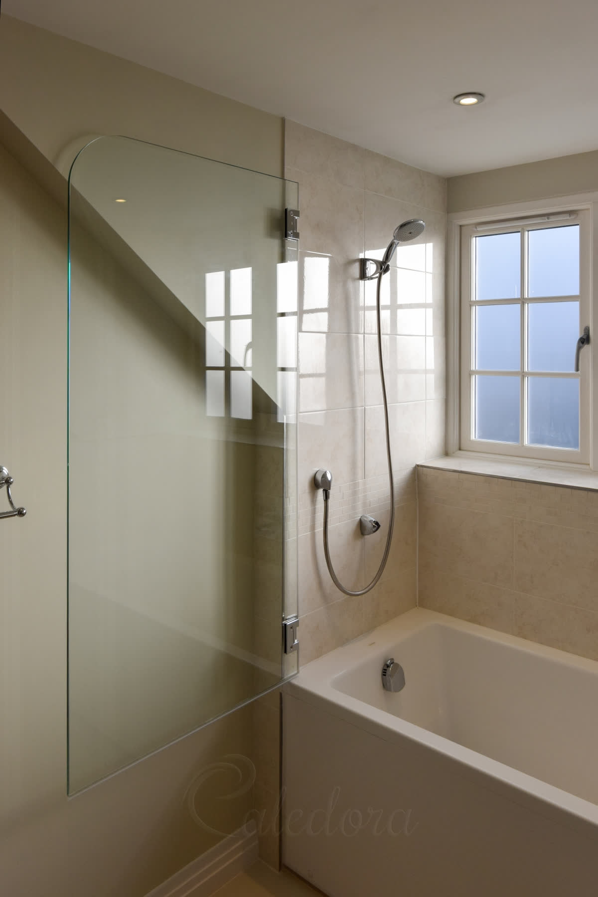 A bath screen with a rounded top corner, hung on chrome hinges beside a window, over a white bath with cream tiles