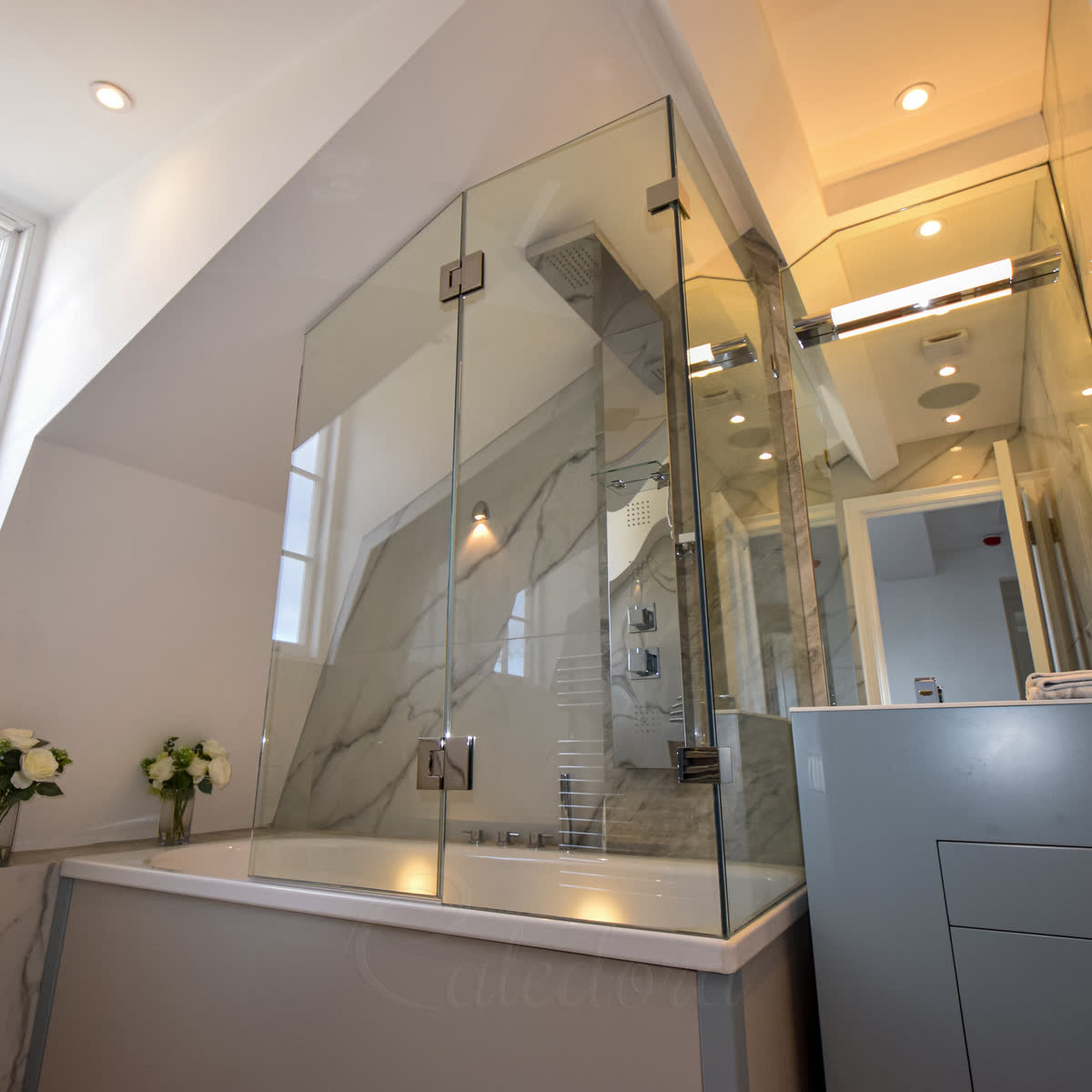 Clear glass on the two open sides of a bath under a roof slope, joined glass to glass, with a hinged panel for access and the shower inside