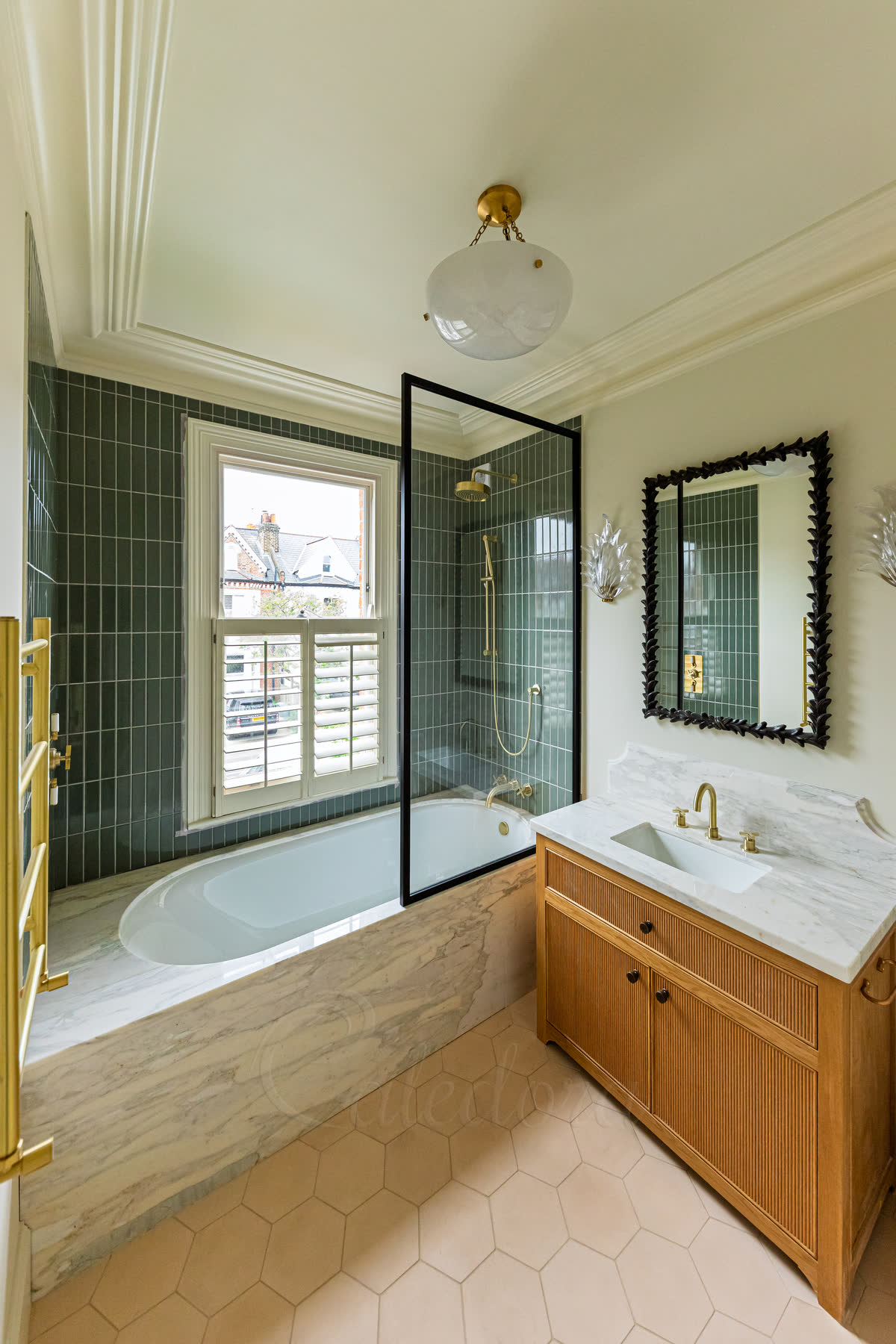 A black-framed fixed bath screen over a marble-panelled bath with green tiles, a brass towel rail and an oak vanity beside it