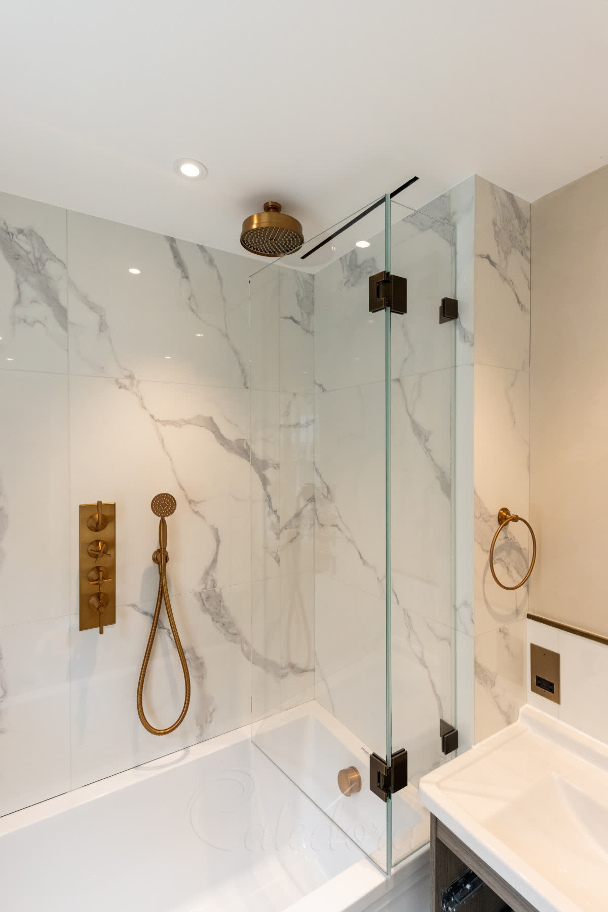 A bath screen with a fixed panel and a hinged panel on antique brass hinges, with white marble walls and a brushed brass rain shower and controls