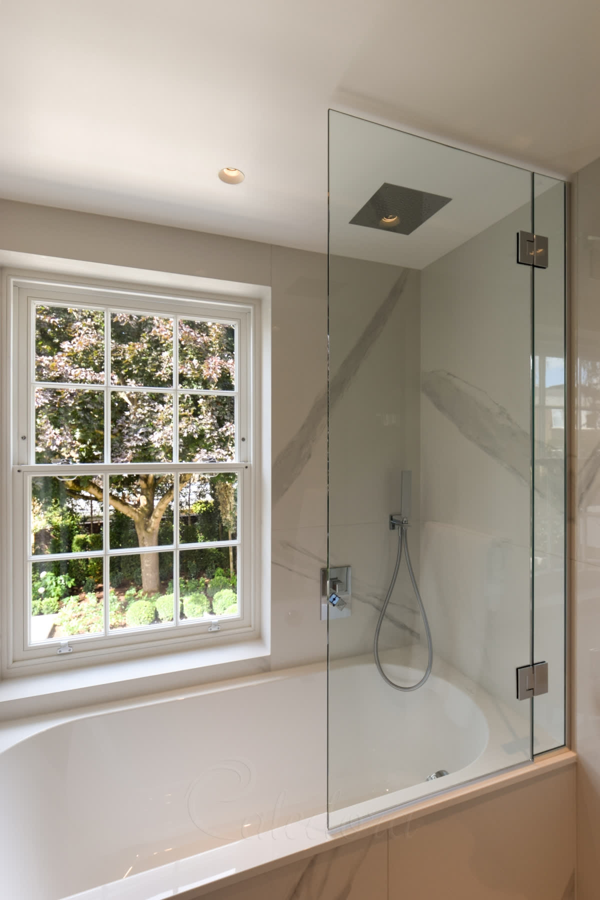 A frameless bath screen with a fixed panel and a hinged panel on polished chrome glass-to-glass hinges, beside a sash window in a white marble bathroom