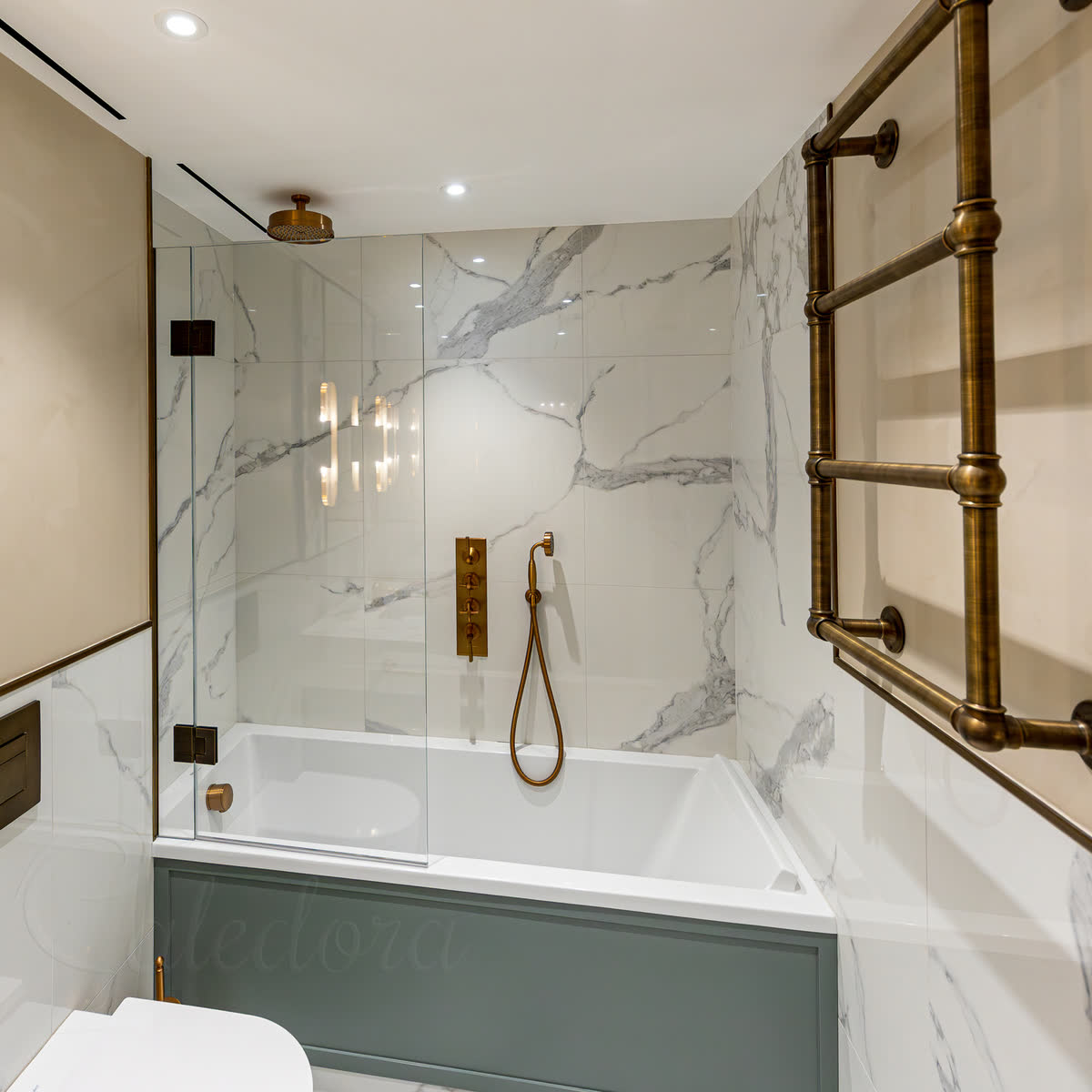 A bath screen with a fixed glass panel at the wall and a hinged panel hung from it on dark hinges, over a white bath in a marble-tiled bathroom with an antique brass shower and towel rail