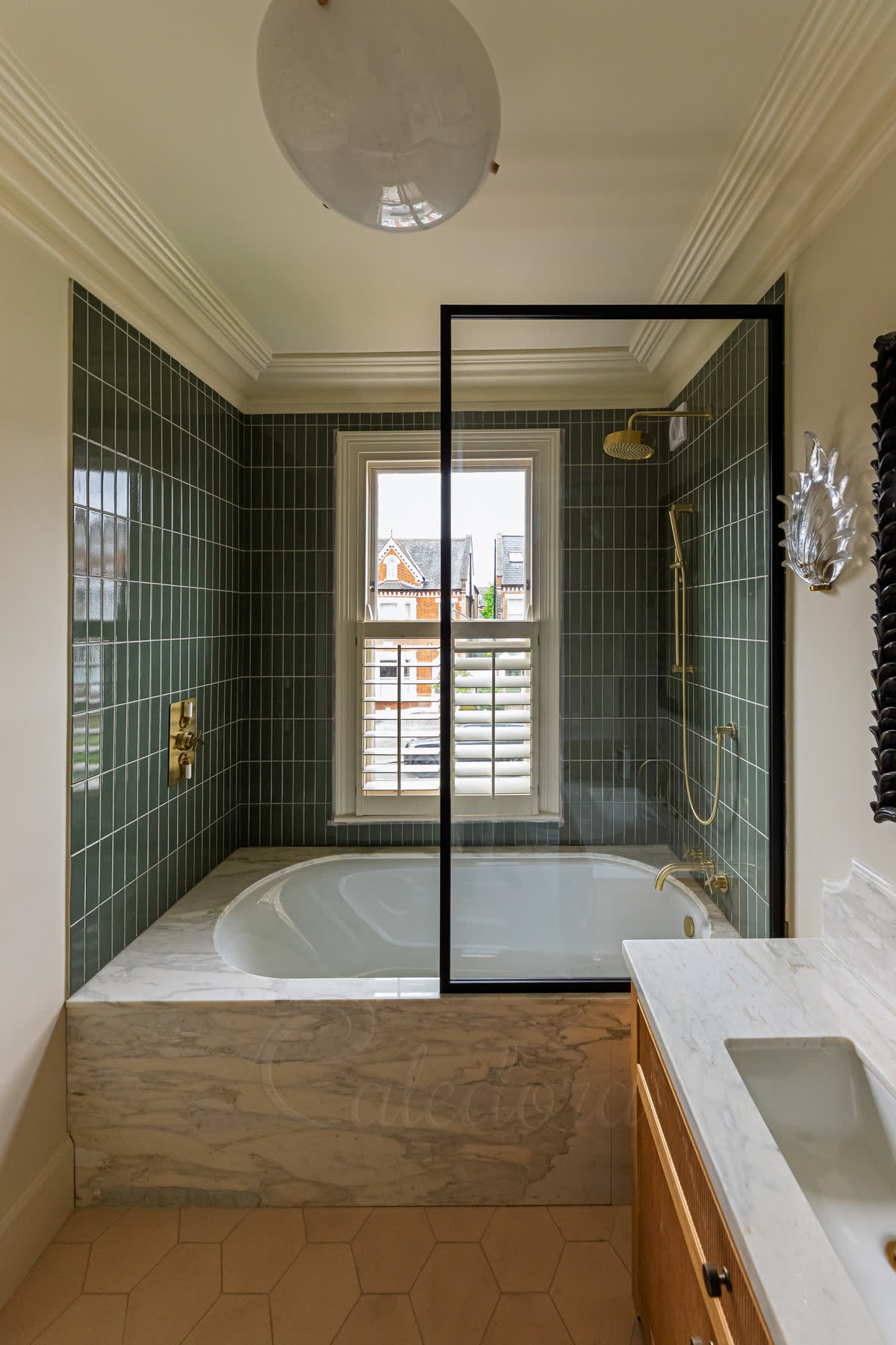 A fixed bath screen in a slim black frame standing on a marble-panelled bath, in front of a sash window and green tiles, with a brass shower behind the glass