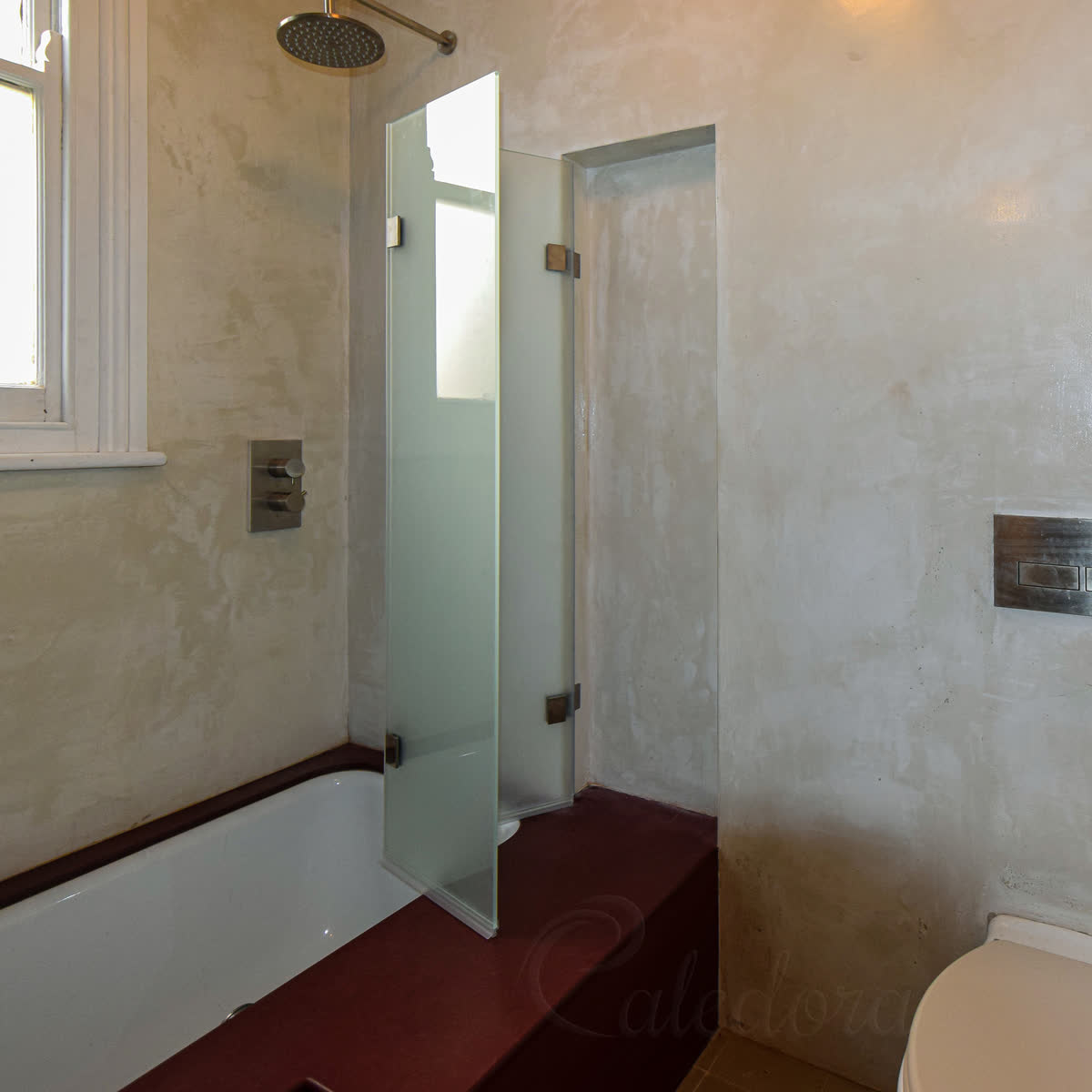 Two frosted glass panels of a folding bath screen opening out from a recess built into the wall, over a bath set in a deep red stone surround