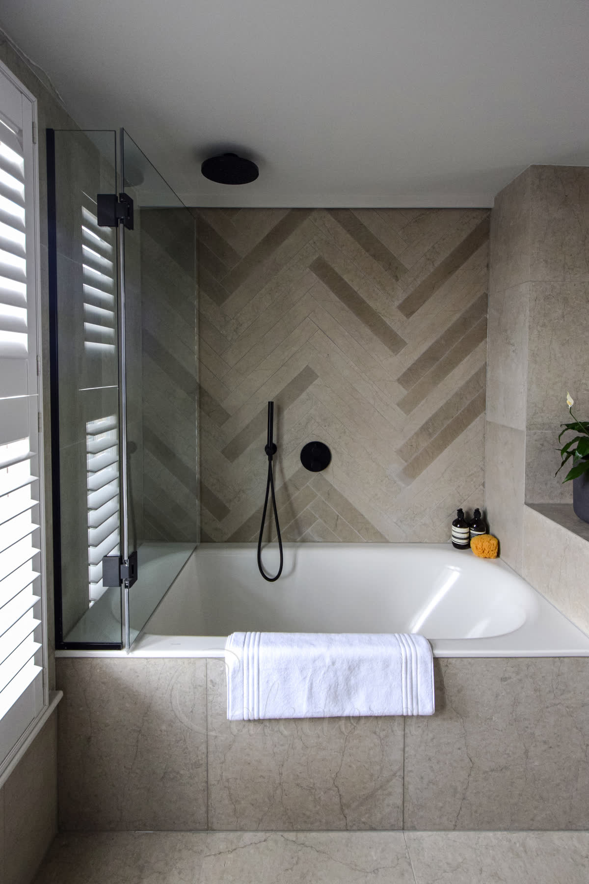 A frameless bath screen on matte black hinges over a bath, against herringbone stone tiles and window shutters