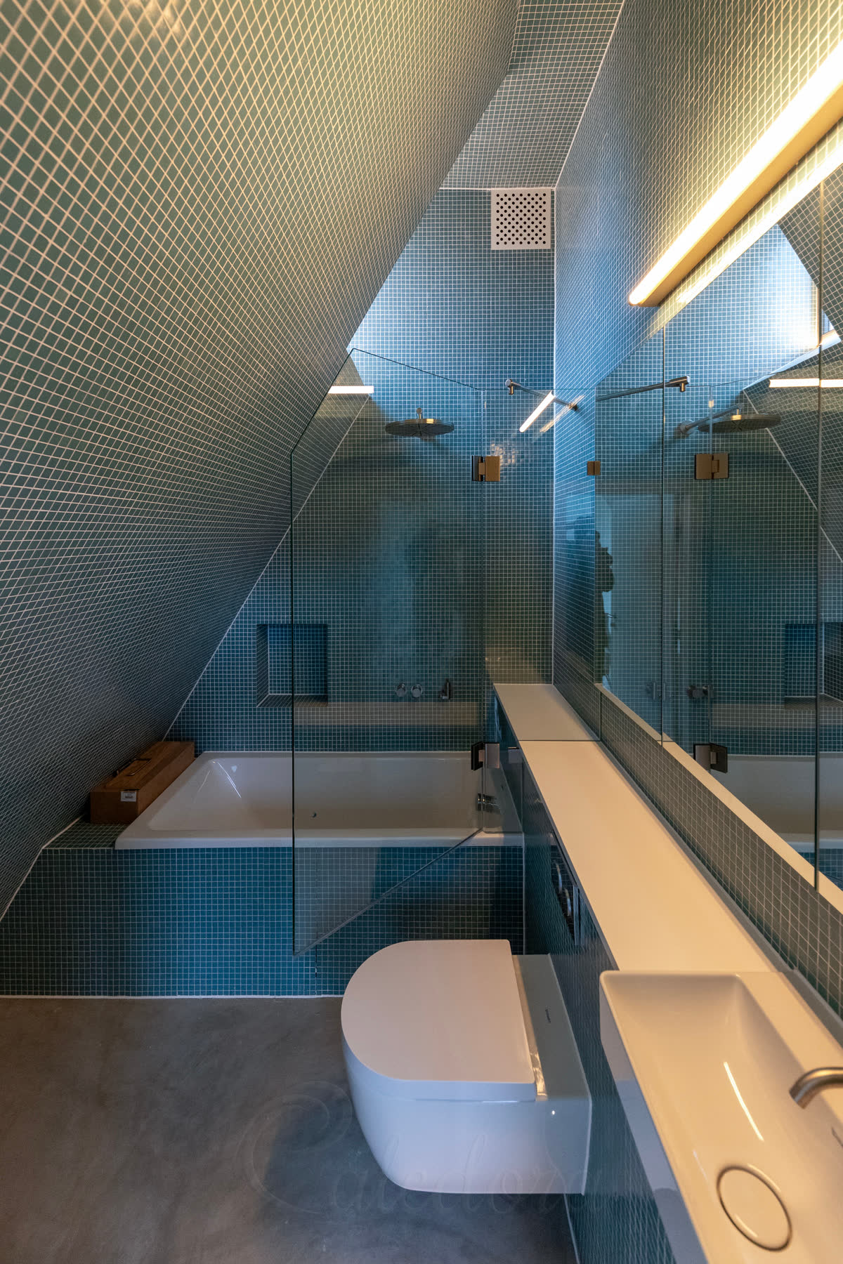 A frameless bath screen under a roof slope in a blue mosaic loft bathroom, its fixed panel notched round the boxing beside the bath