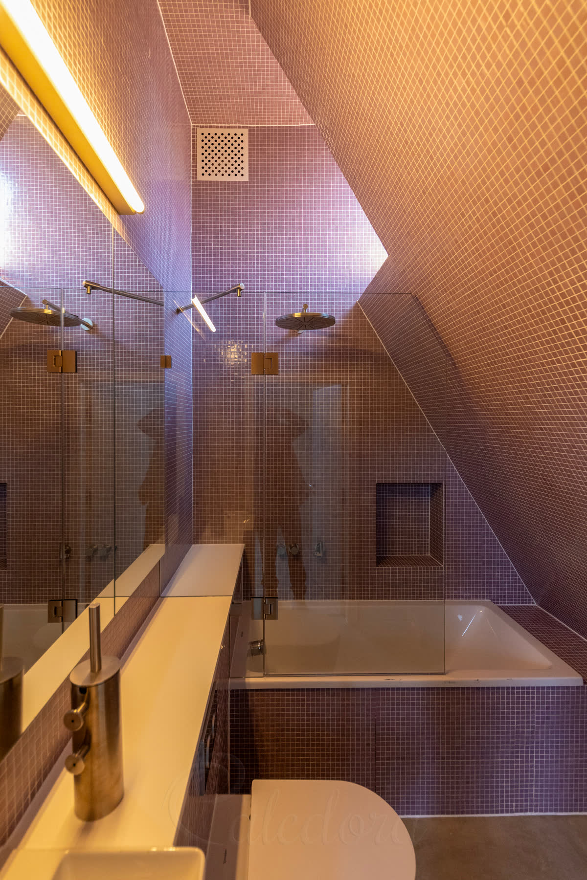 A bath screen made to a loft: the hinged panel cut to the line of the roof slope and the fixed panel notched round the boxing, in a purple mosaic bathroom