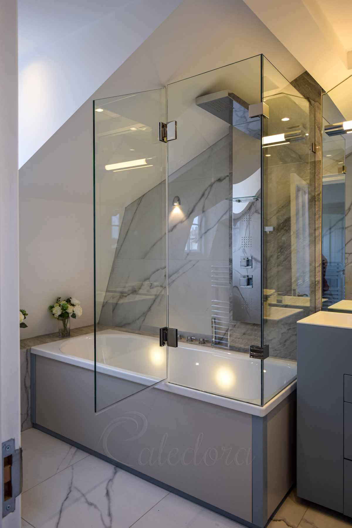 Clear glass on two sides of a bath under a roof slope, joined on polished nickel hinges, with a hinged panel and marble walls behind