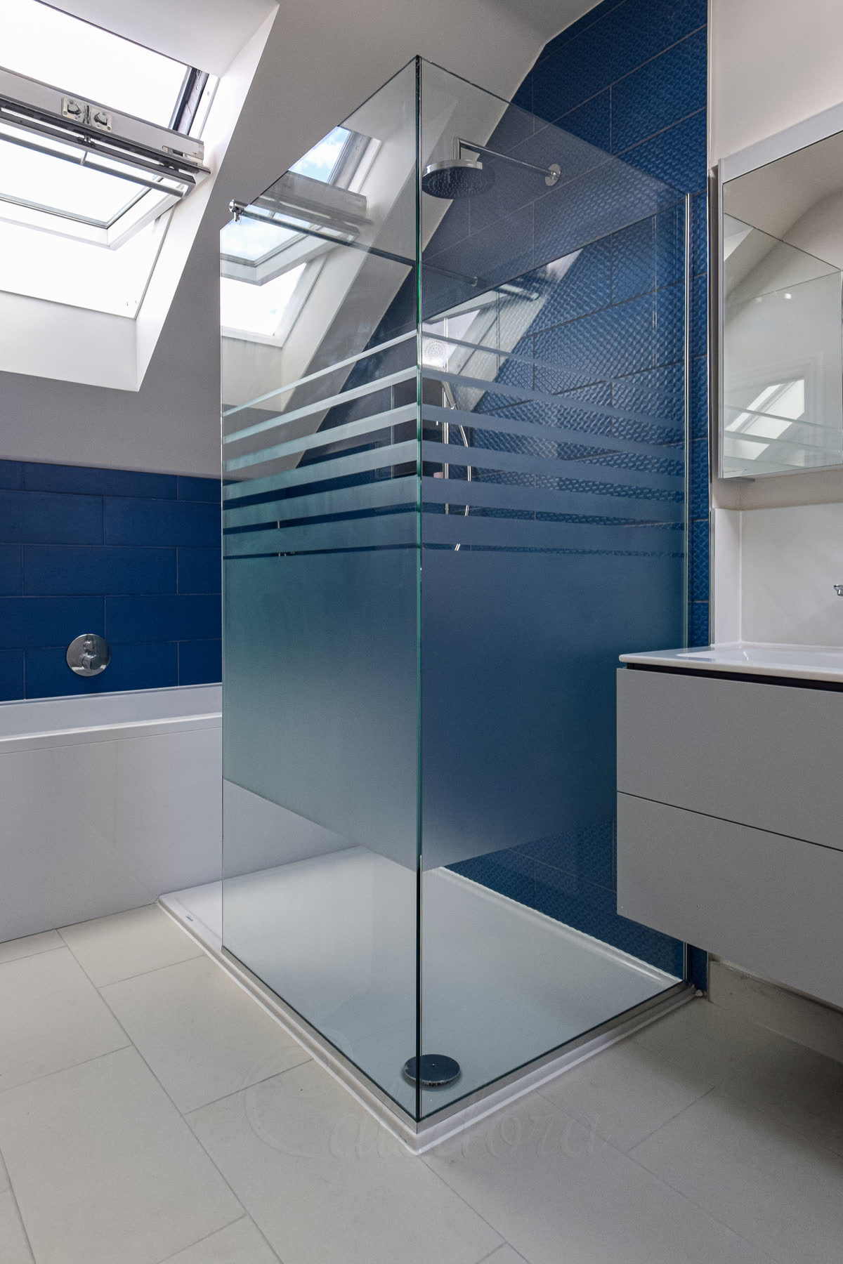 L-shaped corner shower screen with frosted patterned glass for privacy, two fixed panels forming corner enclosure, blue tiles, skylights in loft bathroom