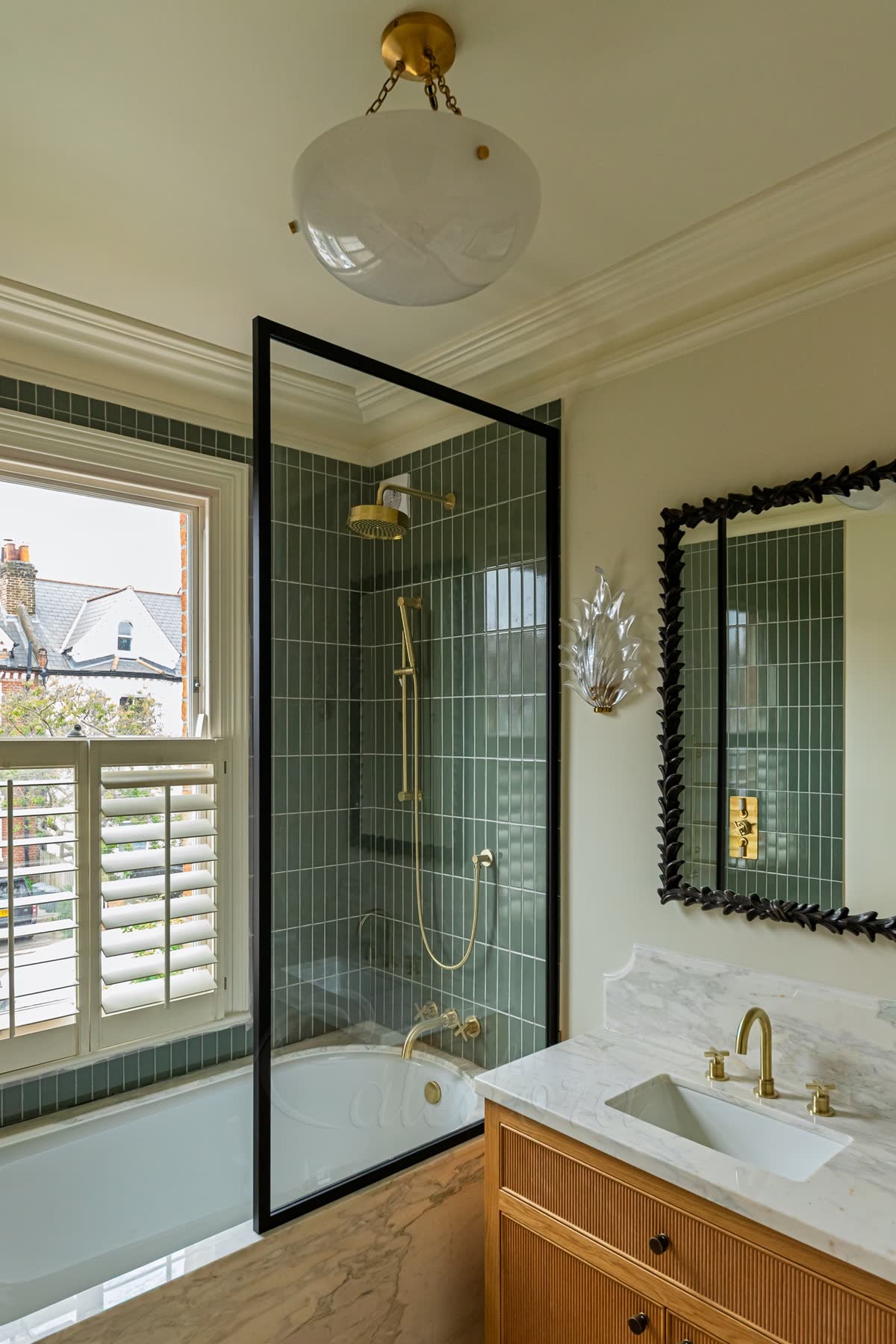 A fixed shower screen on a bath — one black-framed clear glass panel standing on the bath edge, with green vertical tiles, brushed brass shower fittings and a sash window beside it