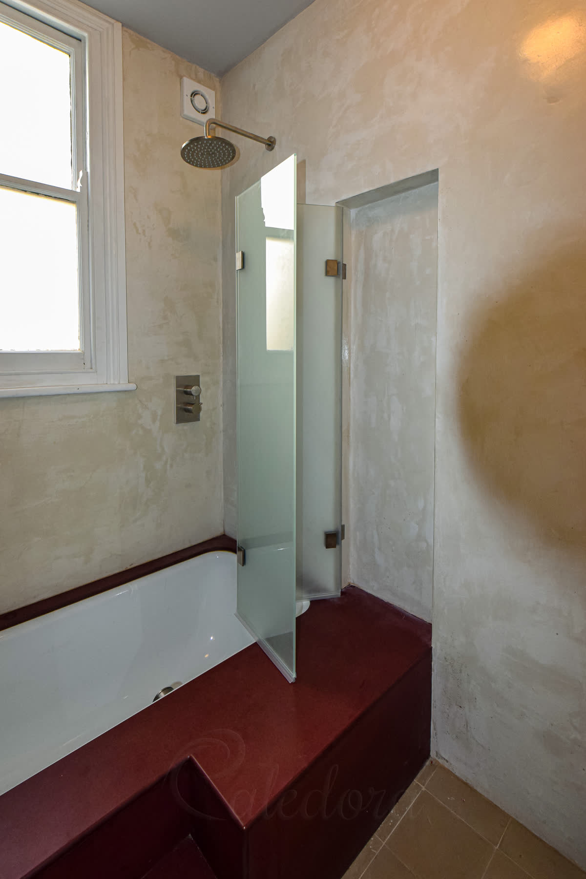 A folding shower screen on a bath, part-folded — two frosted glass panels joined by hinges folding back towards the wall, on a dark red bath surround with plastered walls and a round shower head