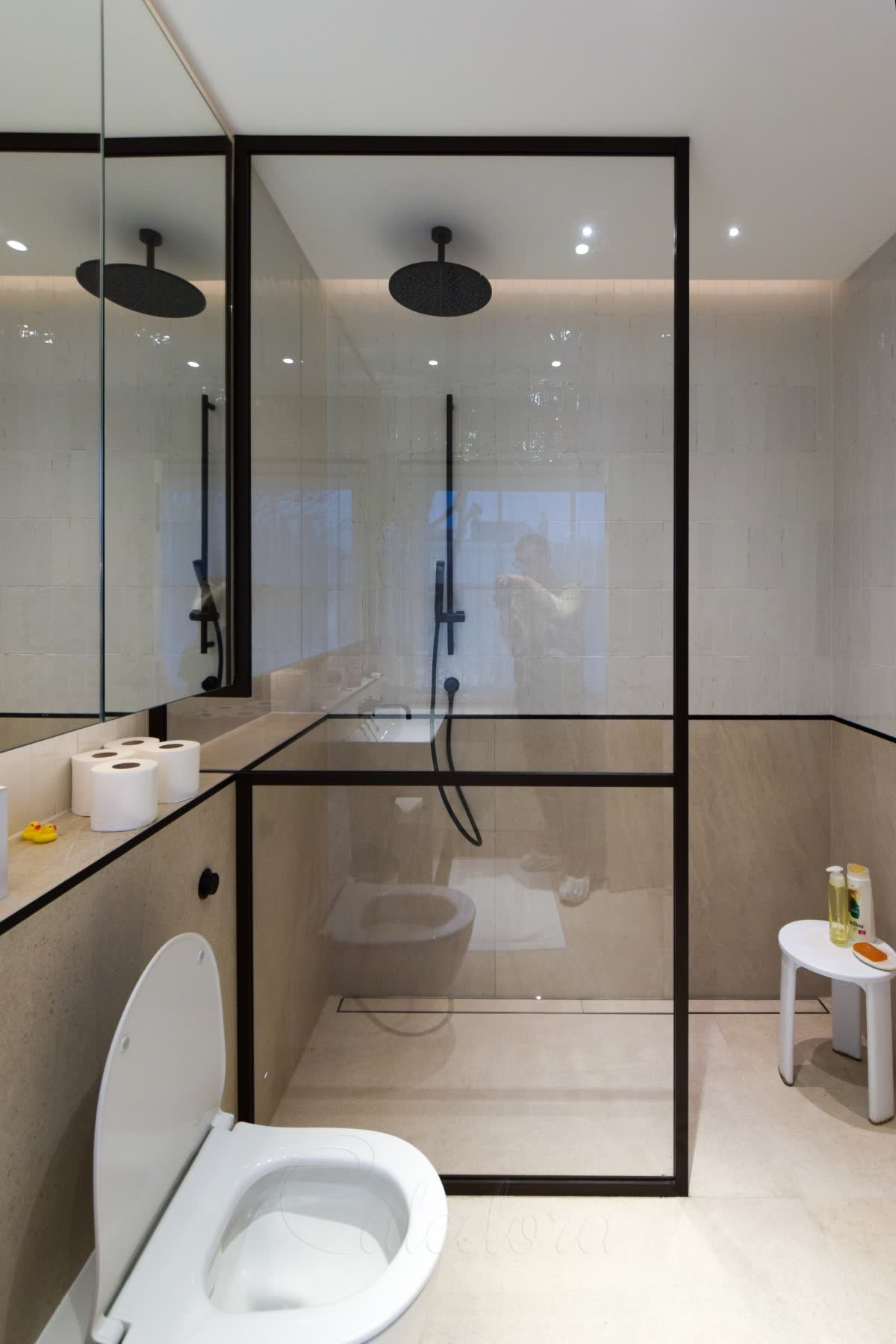 A black-framed walk-in screen rising to the ceiling with one slim crossbar lined up with the stone dado, on a level wet-room floor with a linear drain, white glazed tiles above, stone below and a black ceiling rain head