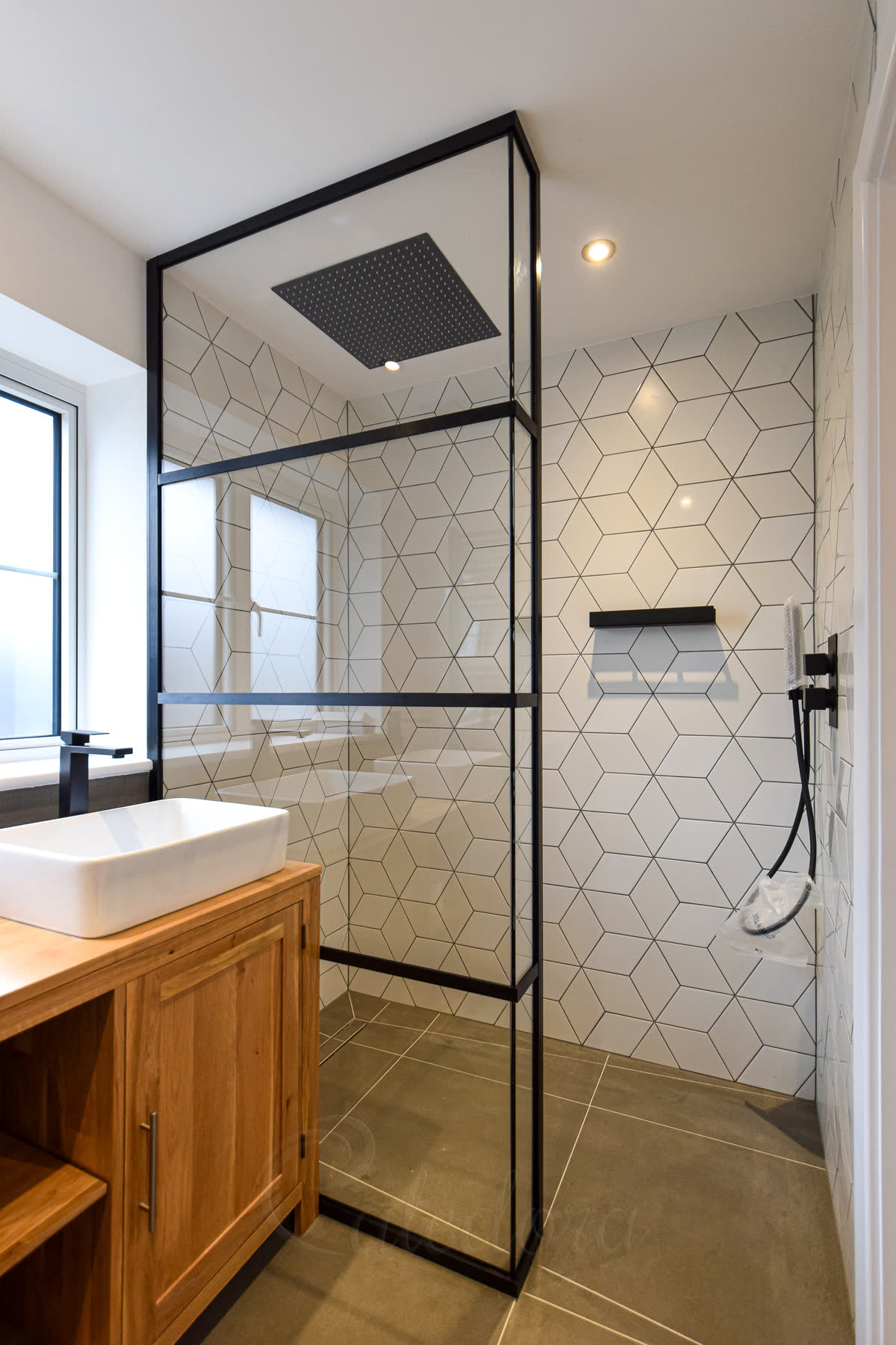 An L-shaped walk-in screen in a black frame with two crossbars, a short return turning in at one end beside the way in, on a grey tiled wet-room floor with a linear drain, white diamond tiles and a black square ceiling rain head