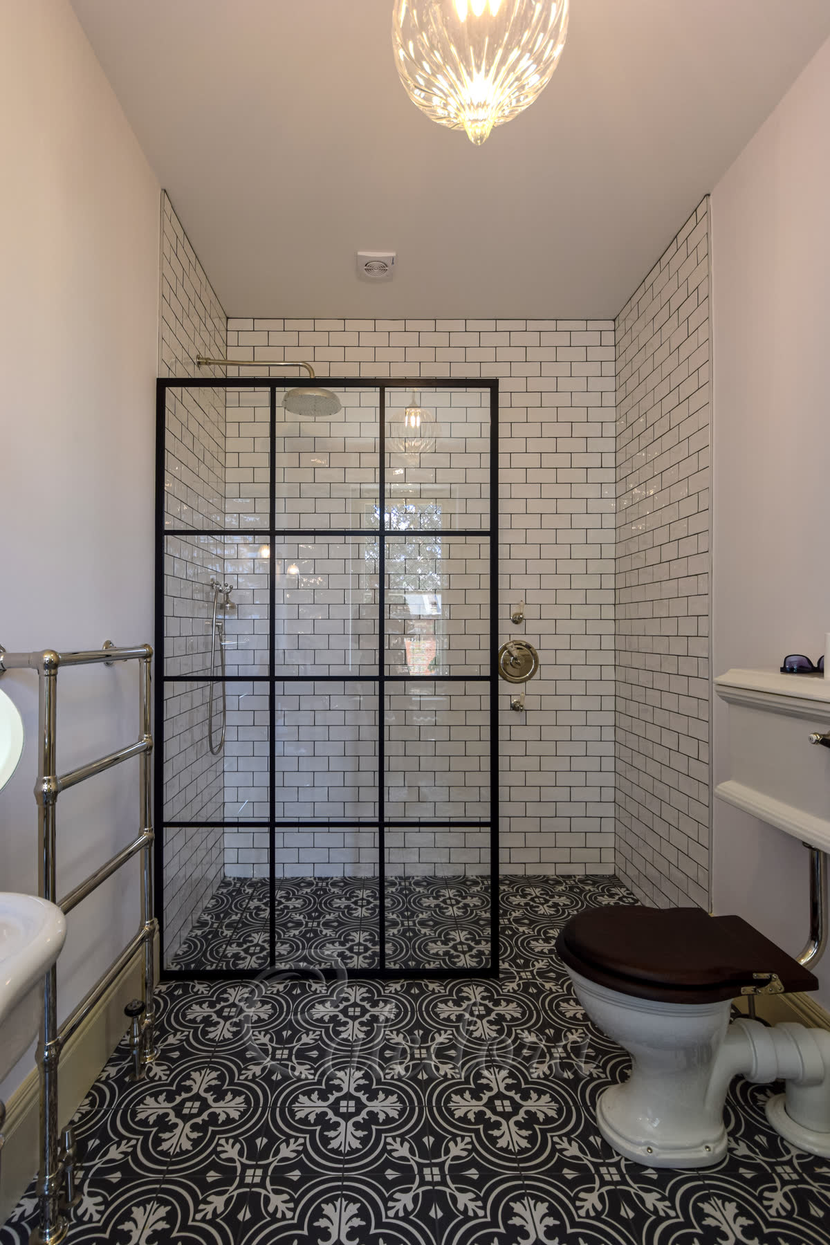 A black grid walk-in screen of twelve panes on a level wet-room floor of black and white patterned tiles that runs on into the bathroom, white metro tiles inside and a polished nickel exposed shower