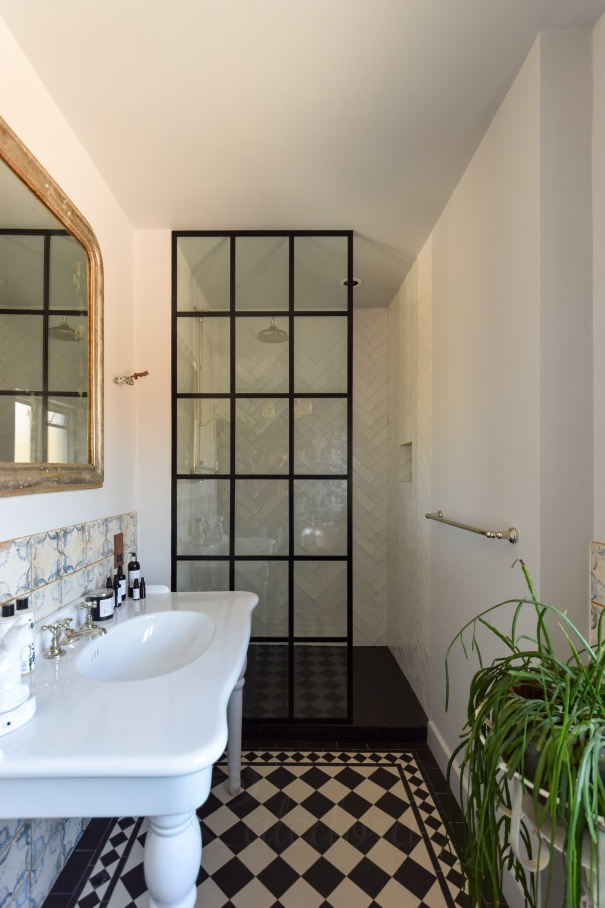 A black grid walk-in screen of fifteen panes standing on a raised black mosaic shower floor, white herringbone tiles inside, with a black and white chequered Victorian floor, a gilt mirror and a washstand in the room