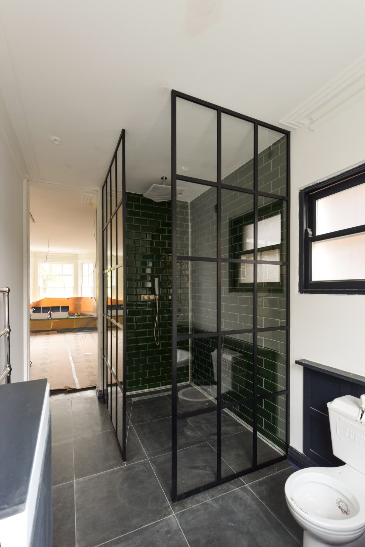 A black grid walk-in shower built from two panels, one along the side and one across the front, with the gap between them left as the way in, on a dark stone wet-room floor with a linear drain and bottle-green metro tiles inside