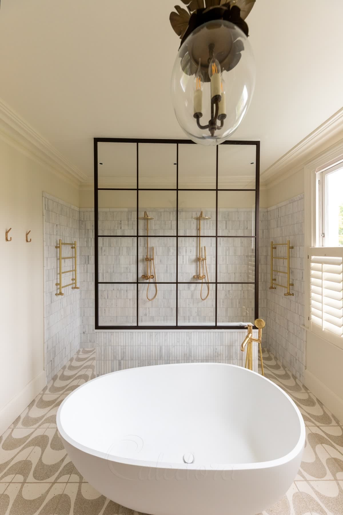 A dark bronze grid walk-in screen running the full width of the room on a tiled half-wall, with two brushed brass showers side by side behind it, white marble stick tiles, and a freestanding bath with a brass bath filler in front