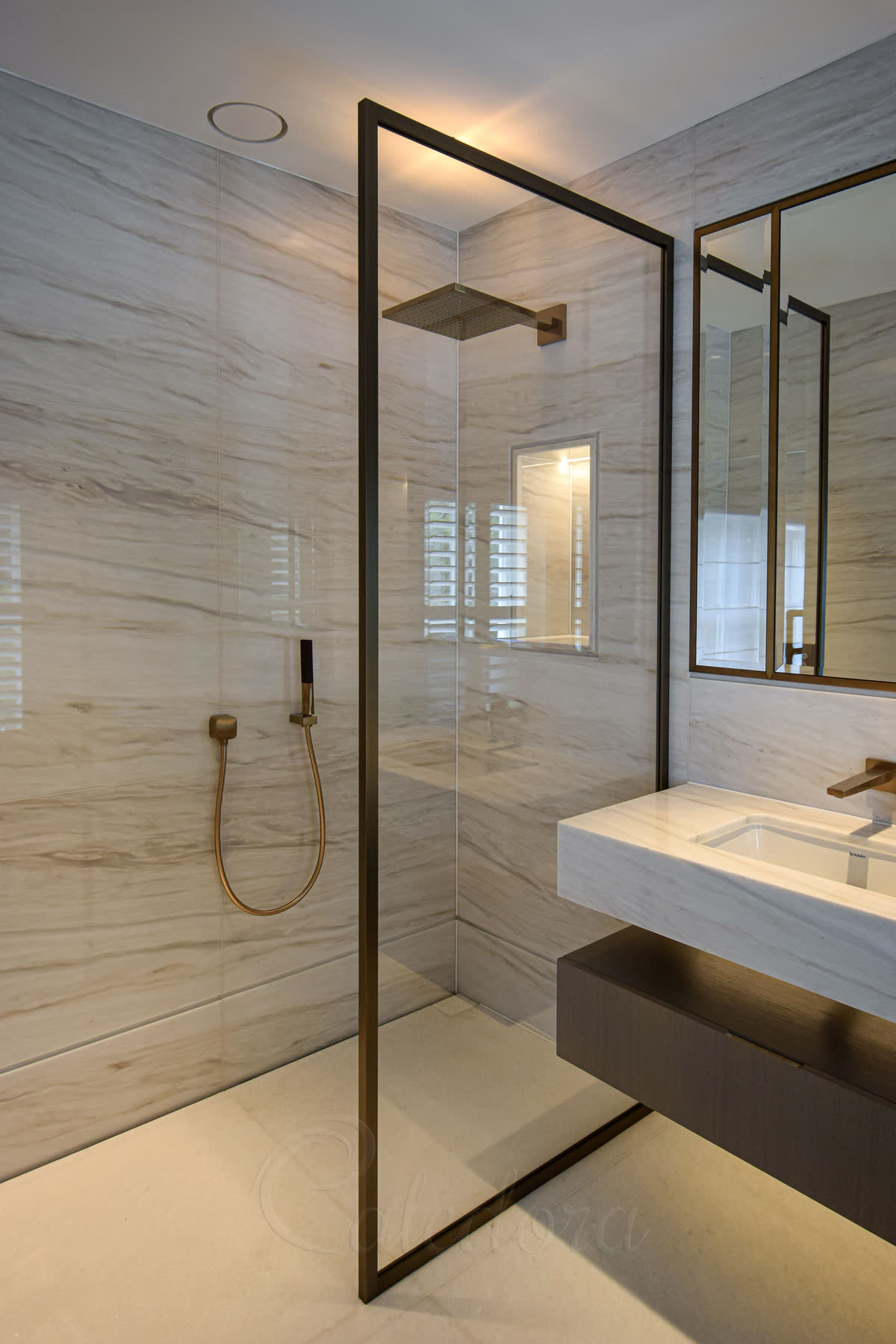 A walk-in panel in a slim brushed bronze frame on a level stone wet-room floor, with a matching bronze shower head, hand shower and basin tap, a lit niche and pale veined marble walls