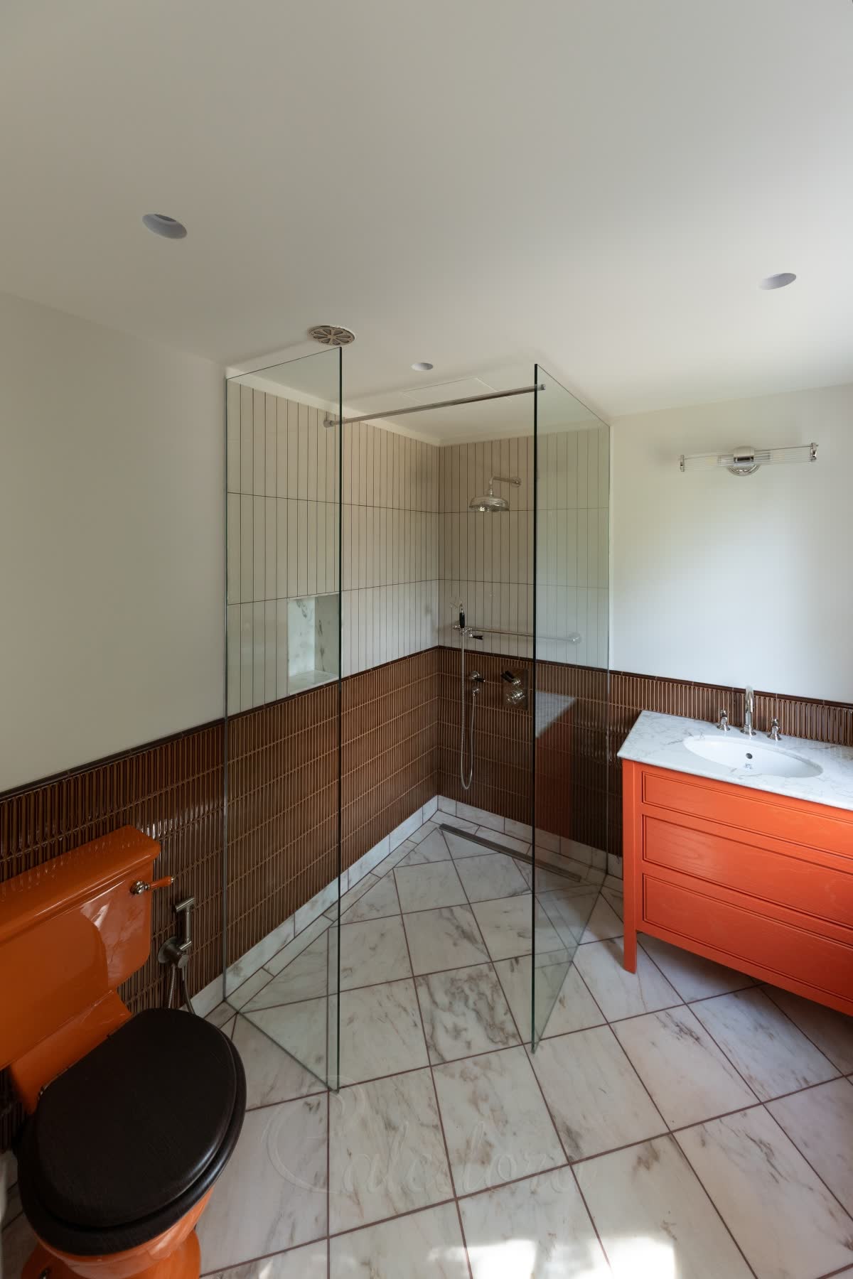 A corner entry walk-in shower — two clear glass panels coming off two walls with the corner between them left open as the way in, a slim bar tying one panel back to the wall, on a level white marble wet-room floor with a linear drain, brown stick tiles and an orange vanity beside it