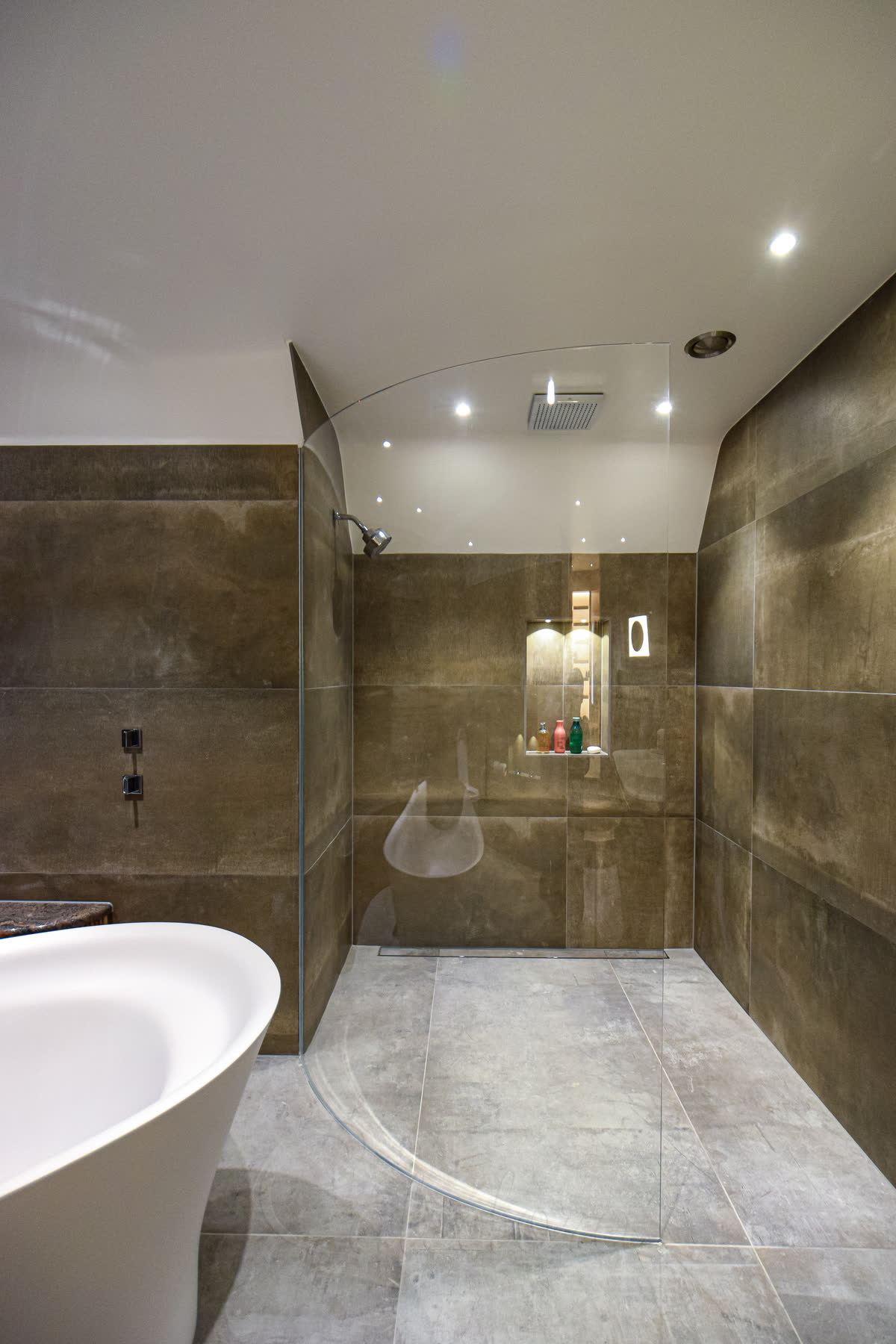 A walk-in screen of curved glass sweeping out from the wall on a level grey stone wet-room floor with a linear drain, deep brown porcelain walls, a lit niche and a freestanding bath beside it