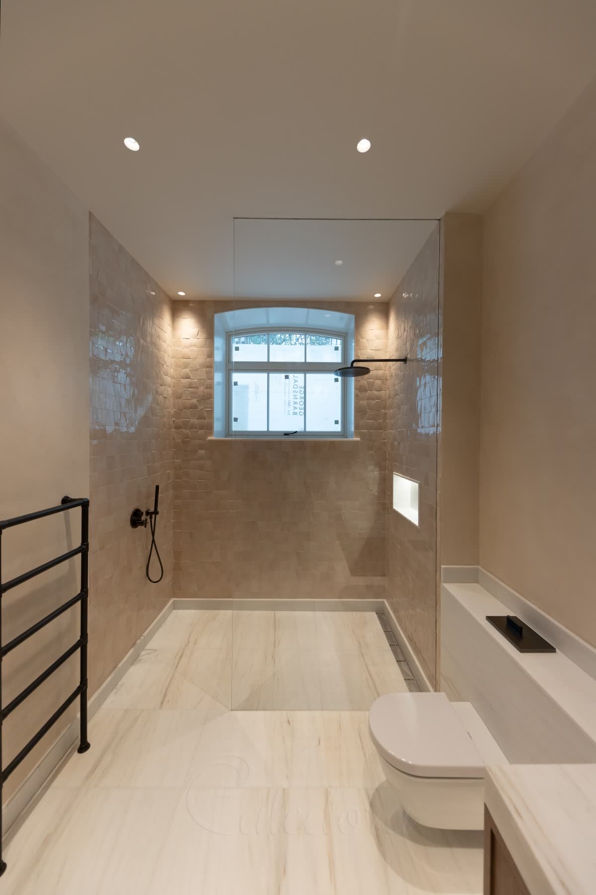 A fixed walk-in shower screen — one plain clear glass panel running up to the ceiling with nothing on it that moves and no visible metal, standing on a level veined stone floor with a linear drain along the wall, under a window in a tiled shower