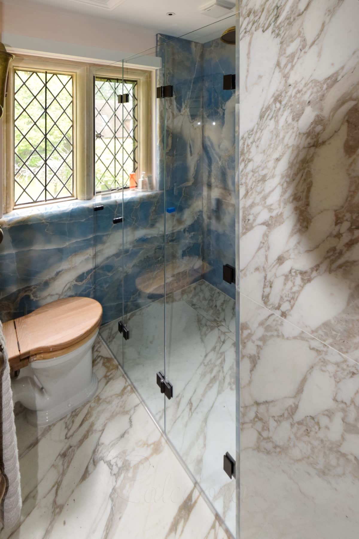 A folding wet room shower screen of three clear glass panels joined by black hinges, closing off a level white and gold veined marble floor with a linear drain along the back wall, blue stone walls inside the shower, a leaded window above and a WC beside it