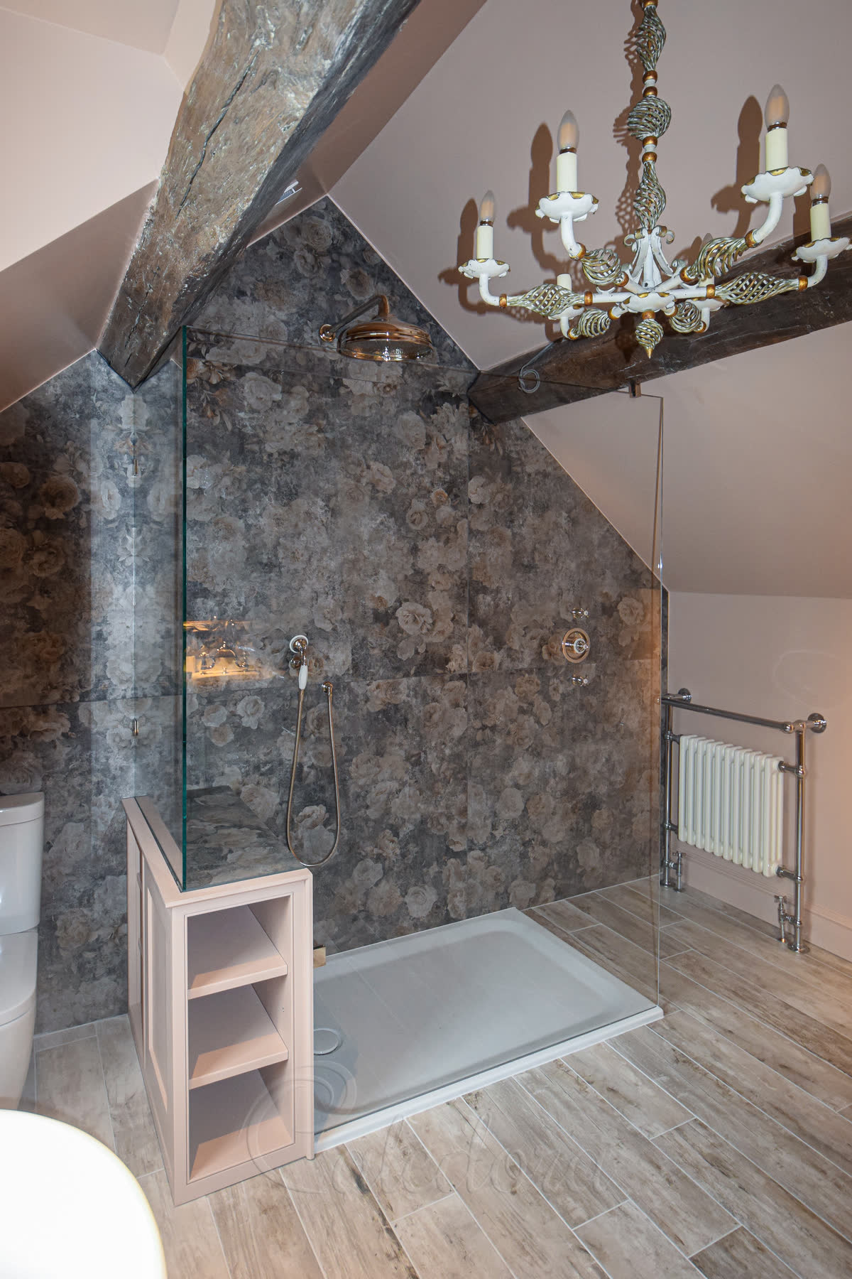 An L-shaped walk-in screen on a white shower tray, its short return standing on top of a painted shelving unit, under a sloping attic ceiling with an old timber beam, grey floral tiles inside and a chandelier overhead